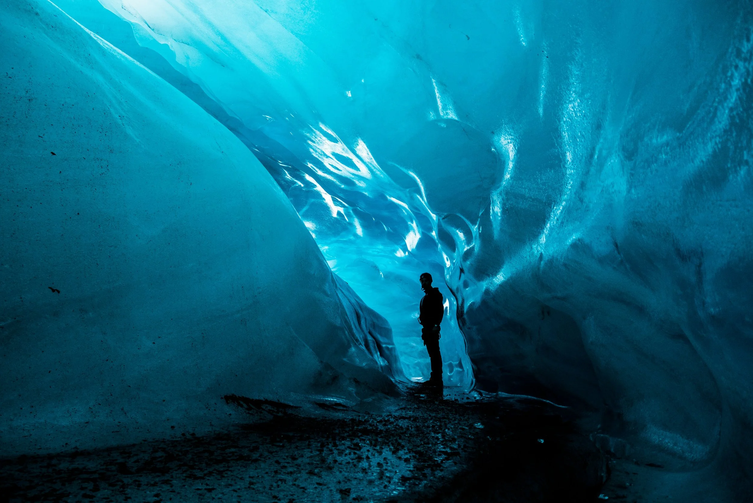 A person silhouetted inside a glowing blue ice cave in the Vatnajökull glacier, surrounded by sculpted translucent walls of ancient glacial ice, Iceland
