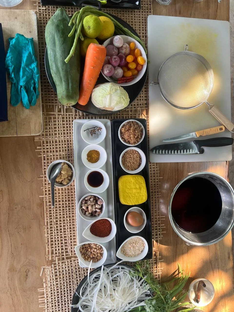 A variety of fresh vegetables, spices, and ingredients arranged on a table for cooking, including zucchini, green beans, carrots, cabbages, cherry tomatoes, shallots, garlic, an egg, turmeric, chili powder, a bowl of soy sauce, and other spices, with