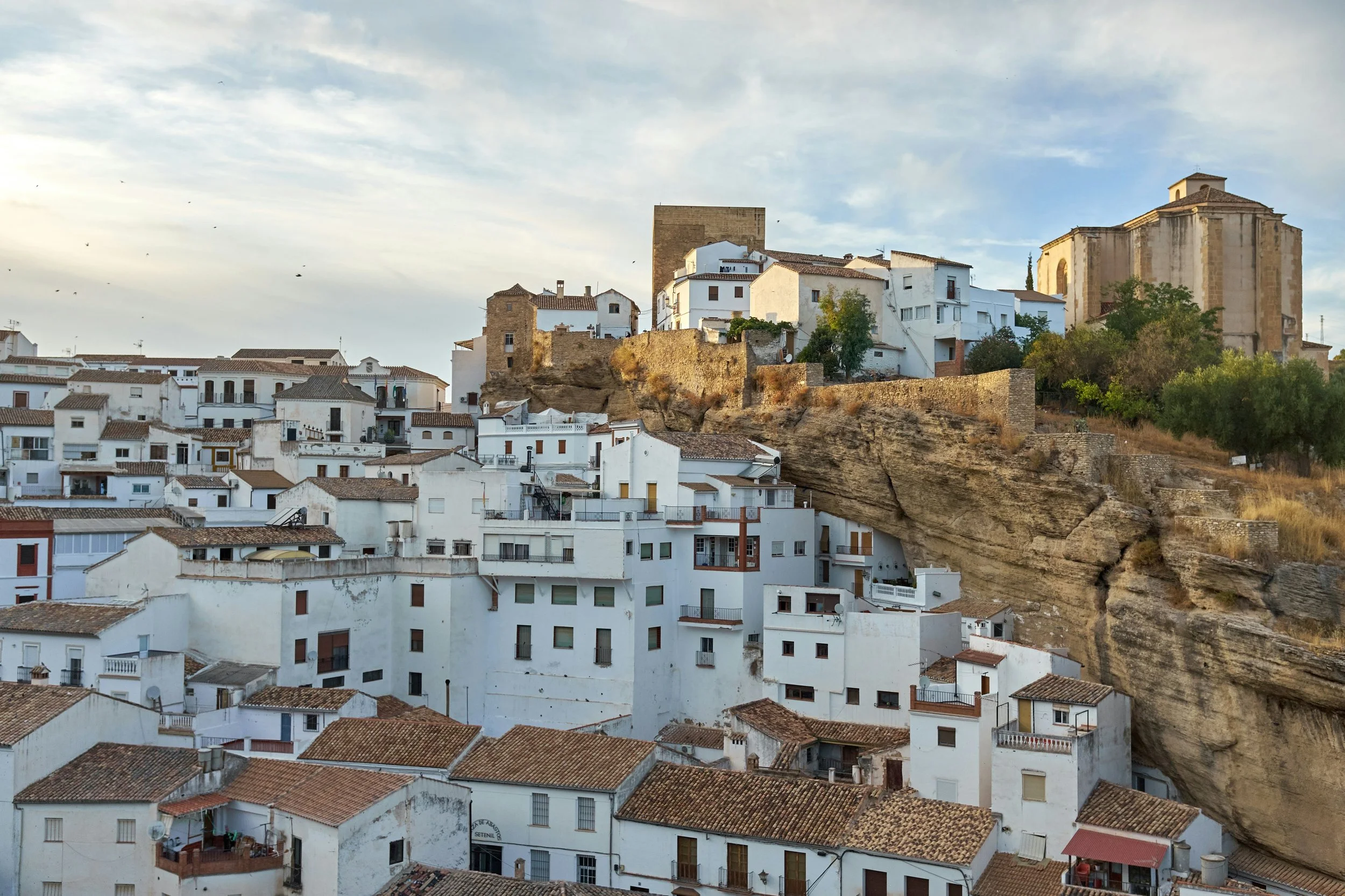 A traditional white-washed Andalusian pueblo blanco clinging to a rocky cliff face under a dramatic cloudy sky, one of Spain's iconic white villages