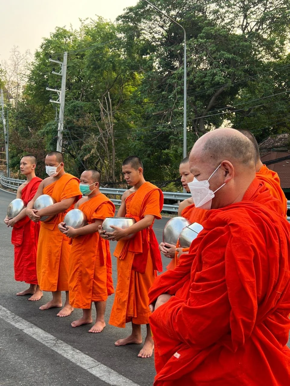 Seven Buddhist monks in orange robes and face masks standing barefoot on the side of a road, holding metal alms bowls, with green trees and utility poles in the background.