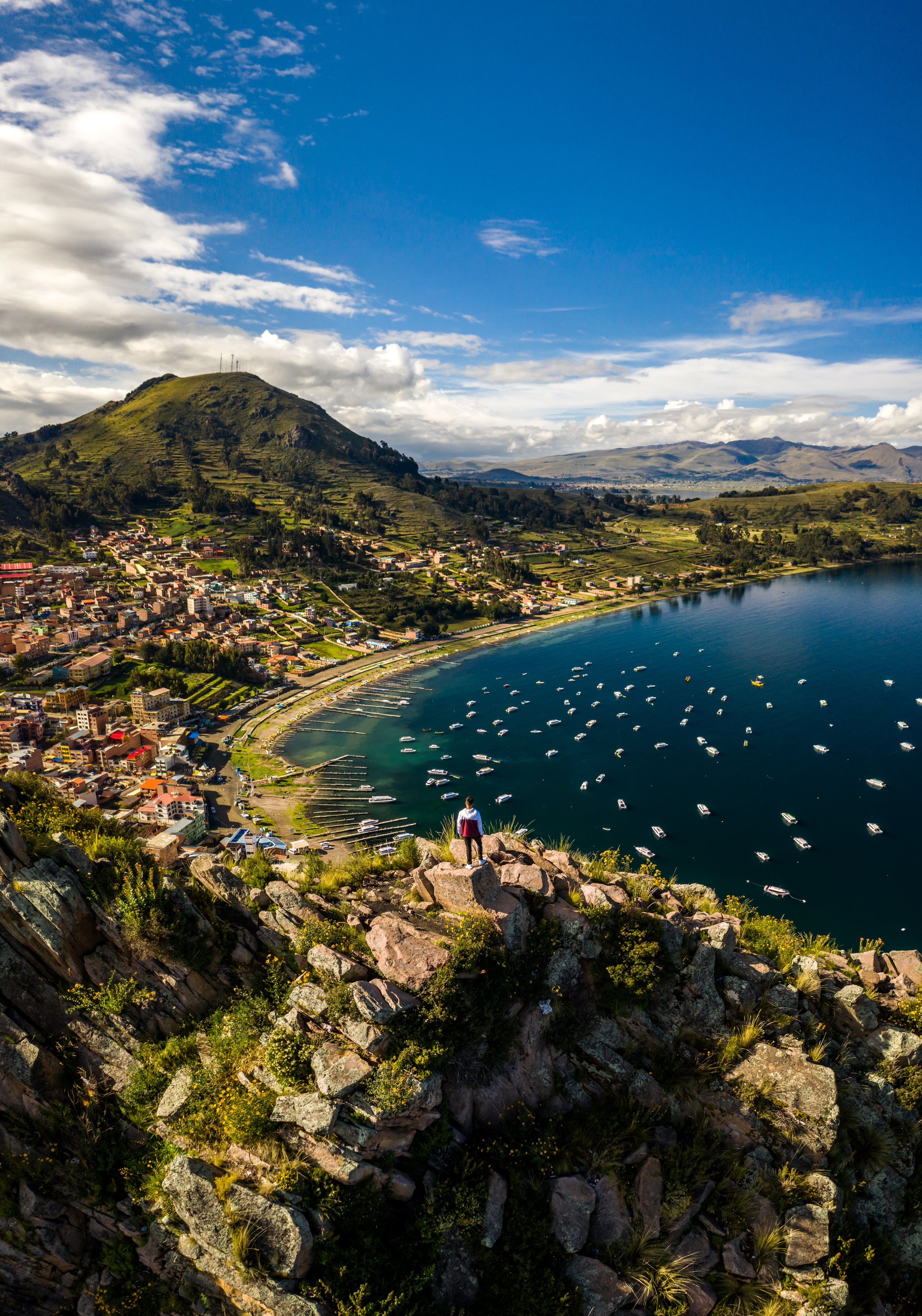 Aerial view of Lake Titicaca with a traveller overlooking a lakeside town, sailboats on deep blue water and a green hill rising behind, Bolivia