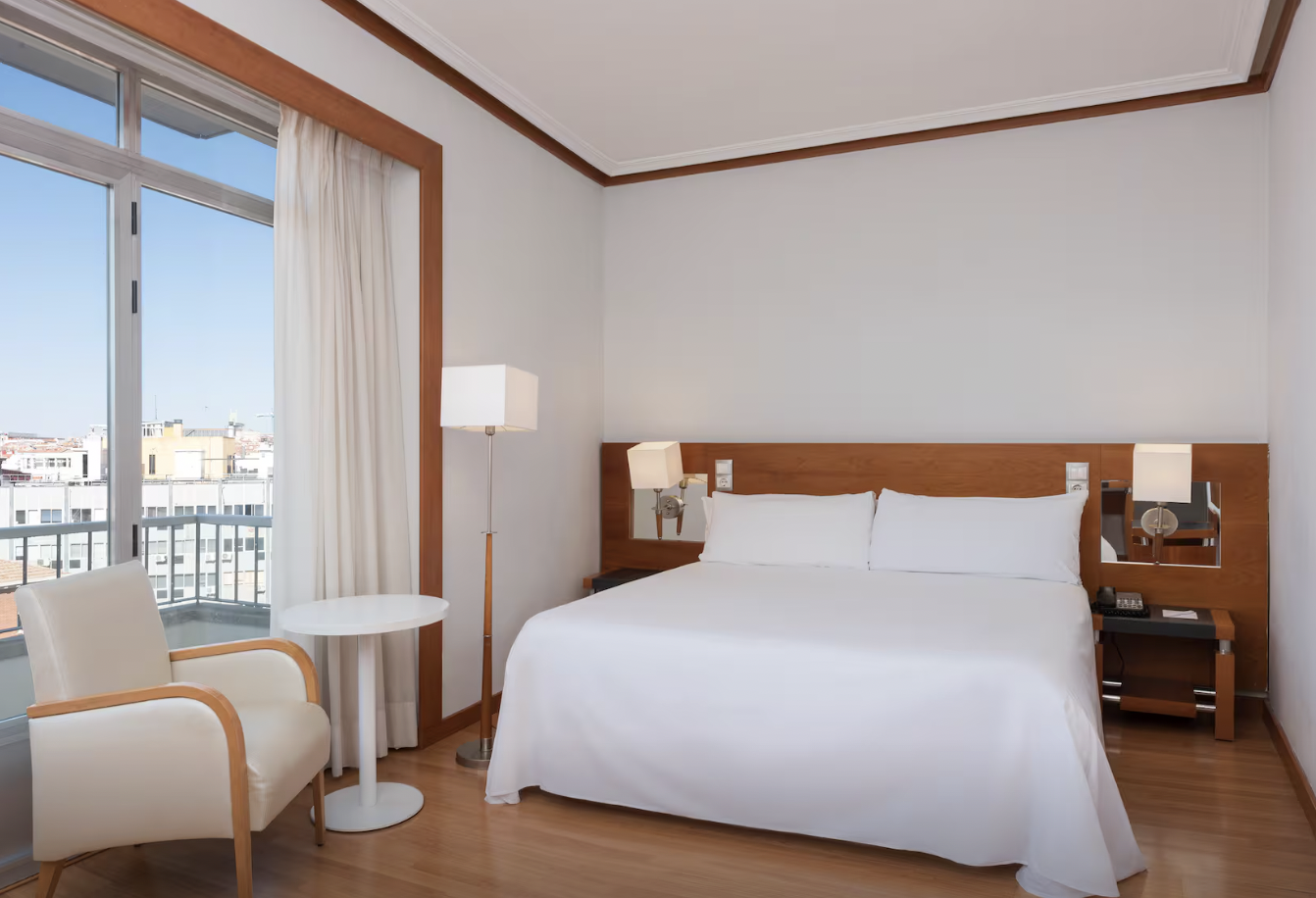 Hotel room with a large bed, bedside lamps, a window with a balcony, an armchair, and a small round table.