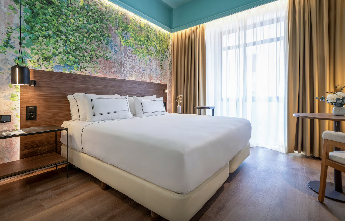 A modern hotel room with a large bed, white bedding, decorative pillows, a wooden headboard, a side table, a hanging black lamp, a wall painting behind the bed, sheer curtains, beige drapes, a small round table, and a chair, with sunlight coming through the large windows.