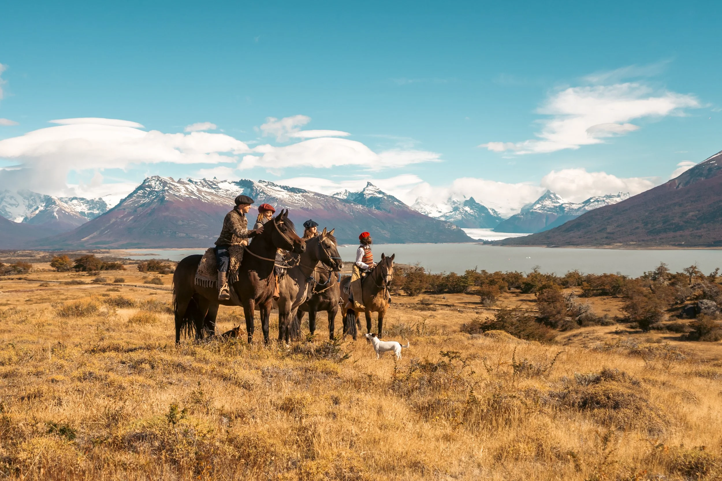 Two riders on horseback crossing golden Patagonian steppe grassland with a glacier and mountain range behind them