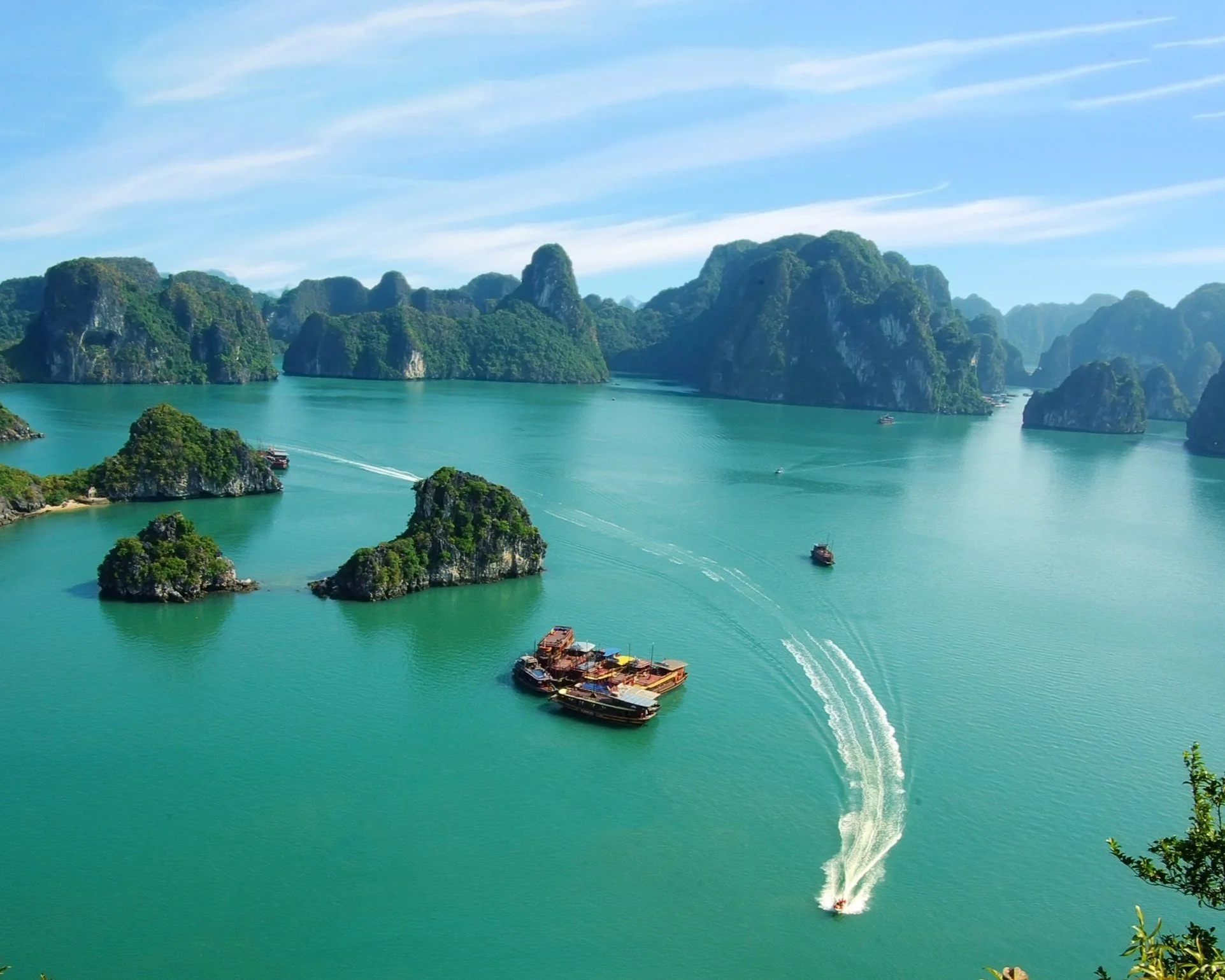 Aerial view of Ha Long Bay in Vietnam showing dozens of forested limestone karst islands rising from jade-green water, with a traditional wooden junk cruise boat cutting a wake across the bay under a blue sky