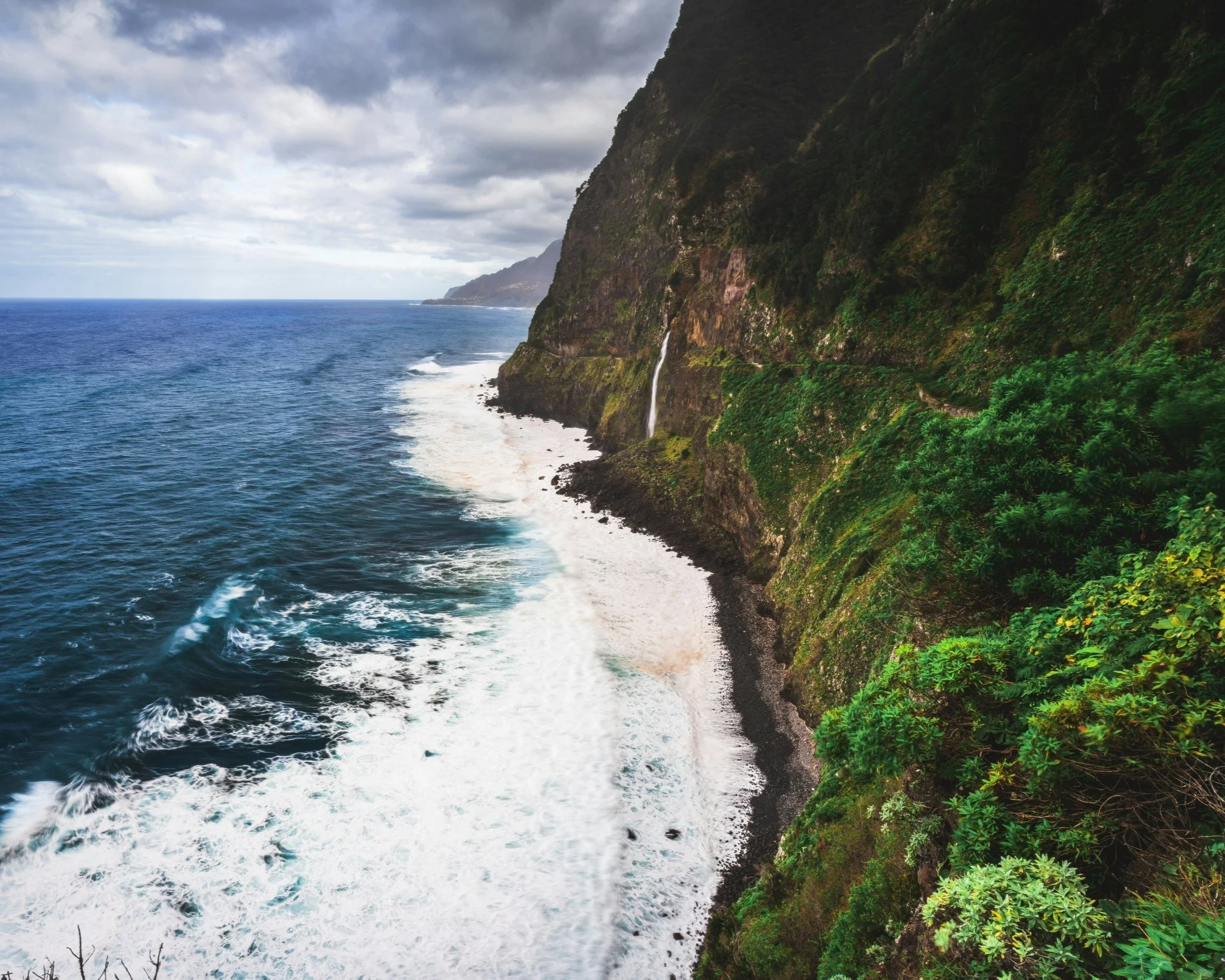 Dramatic green laurisilva-covered cliffs dropping to the Atlantic Ocean along the rugged northern coastline of Madeira, with a waterfall visible on the cliff face, Portugal