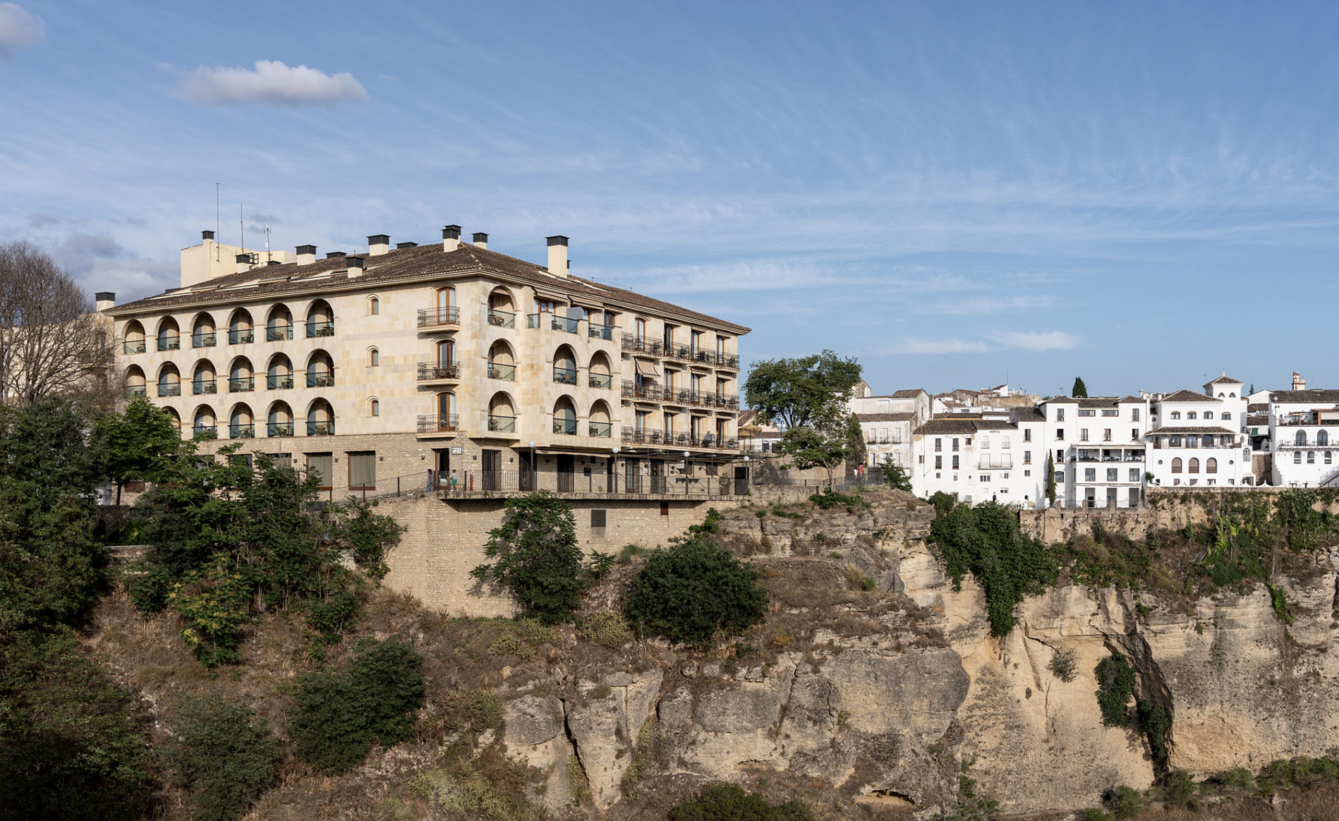 A large beige building with arched windows on a cliffside, overlooking greenery and other white buildings in the background, under a partly cloudy blue sky.