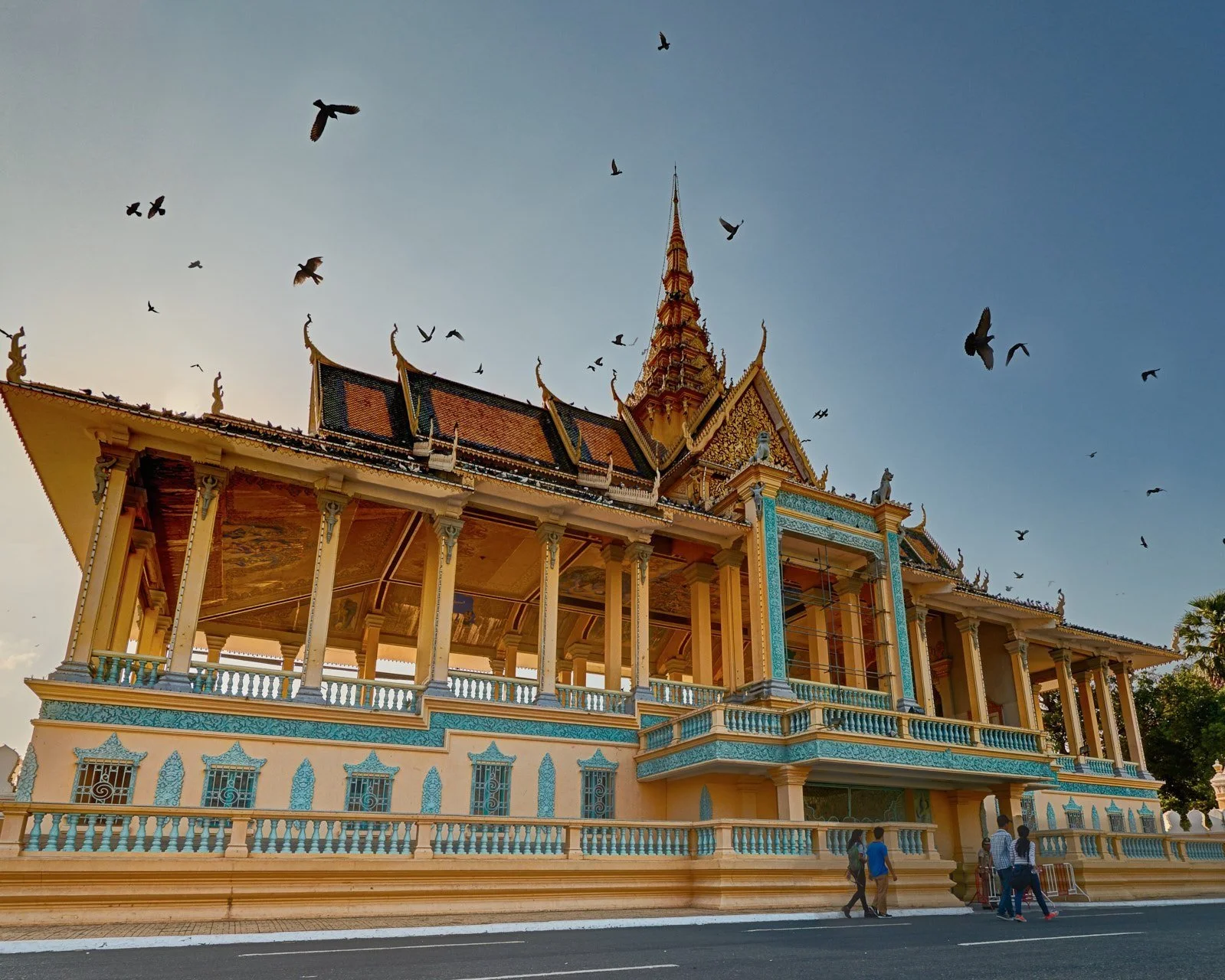 The ornate golden-roofed Silver Pagoda within the Royal Palace complex in Phnom Penh at dusk, with birds flying overhead against a deep blue sky and visitors at the entrance below, Cambodia