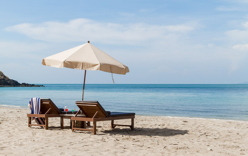 Beach scene with two wooden lounge chairs, an open white umbrella, and a striped towel on one of the chairs, overlooking calm ocean waters under a blue sky with some clouds.