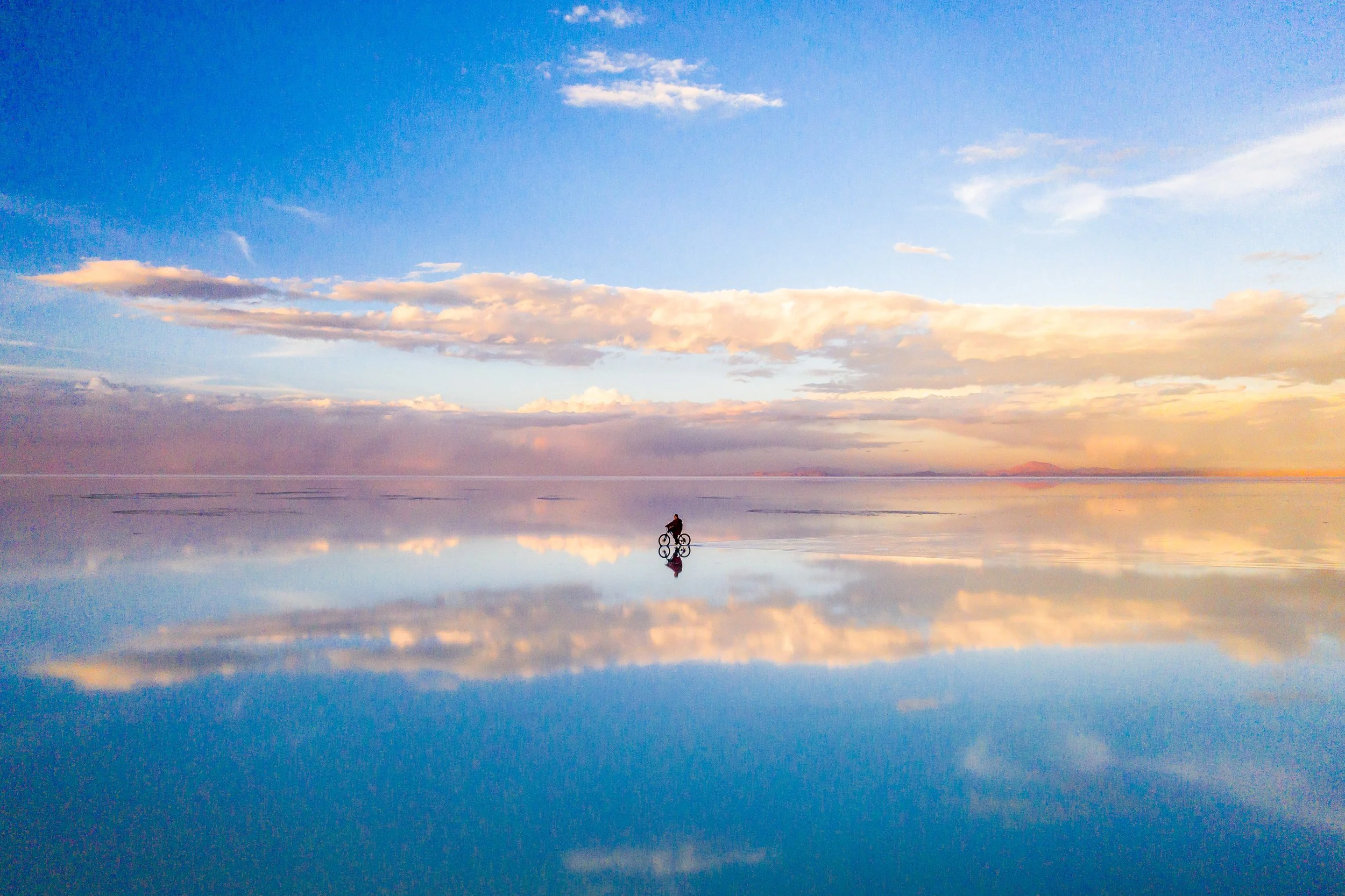 Lone cyclist silhouetted against a golden and blue sunset sky perfectly reflected on the flooded surface of the Salar de Uyuni, Bolivia