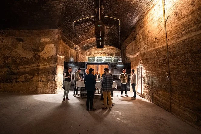 Small group of guests tasting wine inside a centuries-old brick-vaulted underground wine cellar in Hungary, lit by warm ambient light