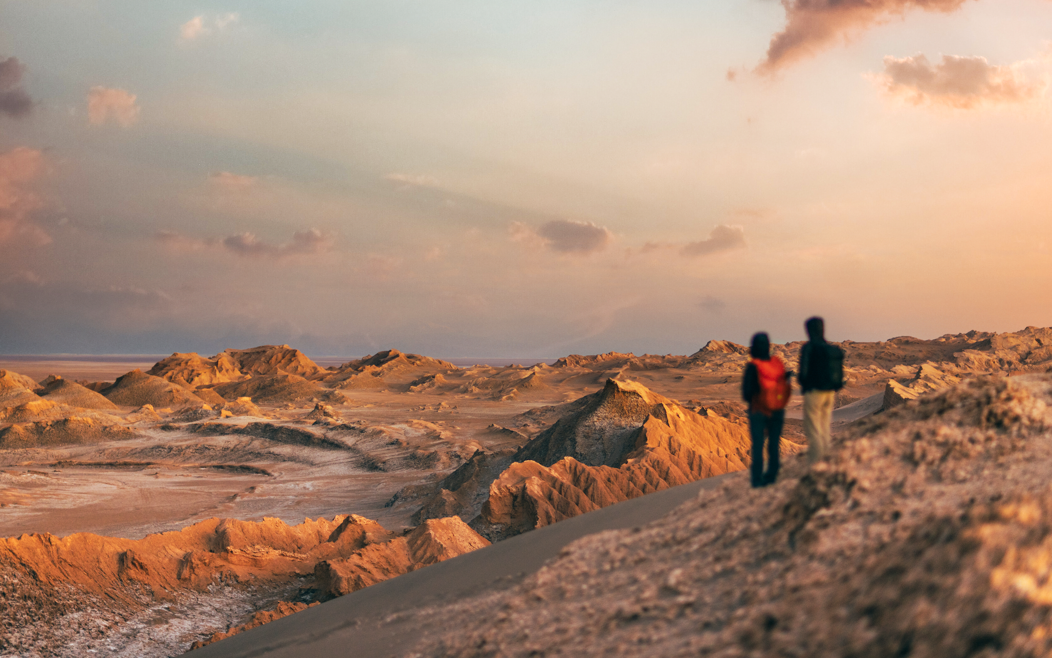 Two hikers walking on desert landscape with rocky formations during sunset.