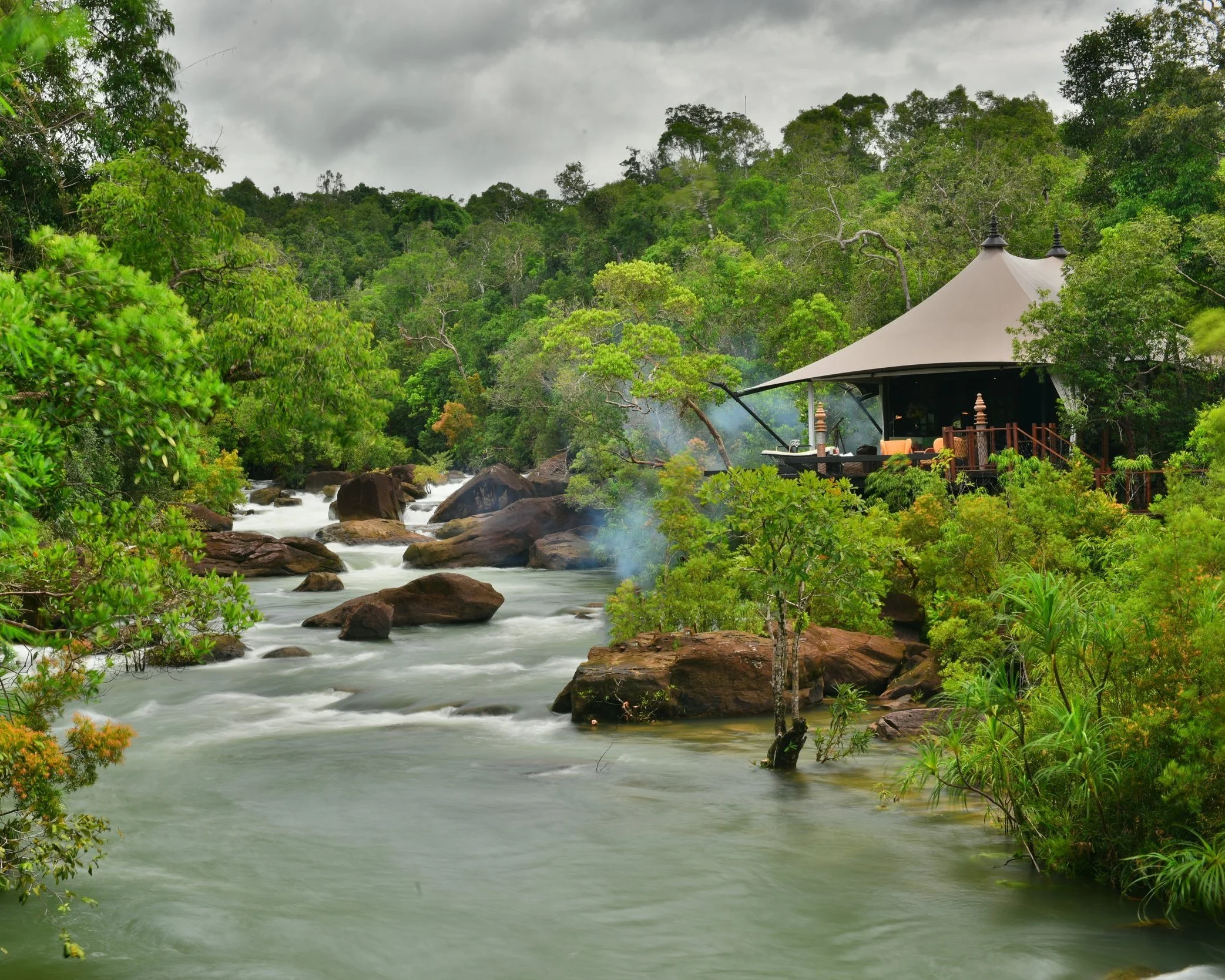 A luxury safari-style tented camp set among tropical forest beside a fast-flowing rocky river in the Cardamom Mountains of Cambodia, with dense jungle canopy surrounding the camp