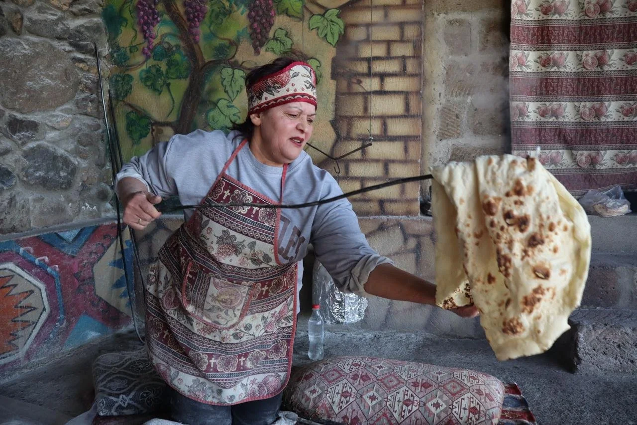 Armenian woman in a traditional embroidered apron pulling a freshly baked sheet of lavash flatbread from a tonir clay oven using a long metal rod, with painted stone and carpet walls visible behind her