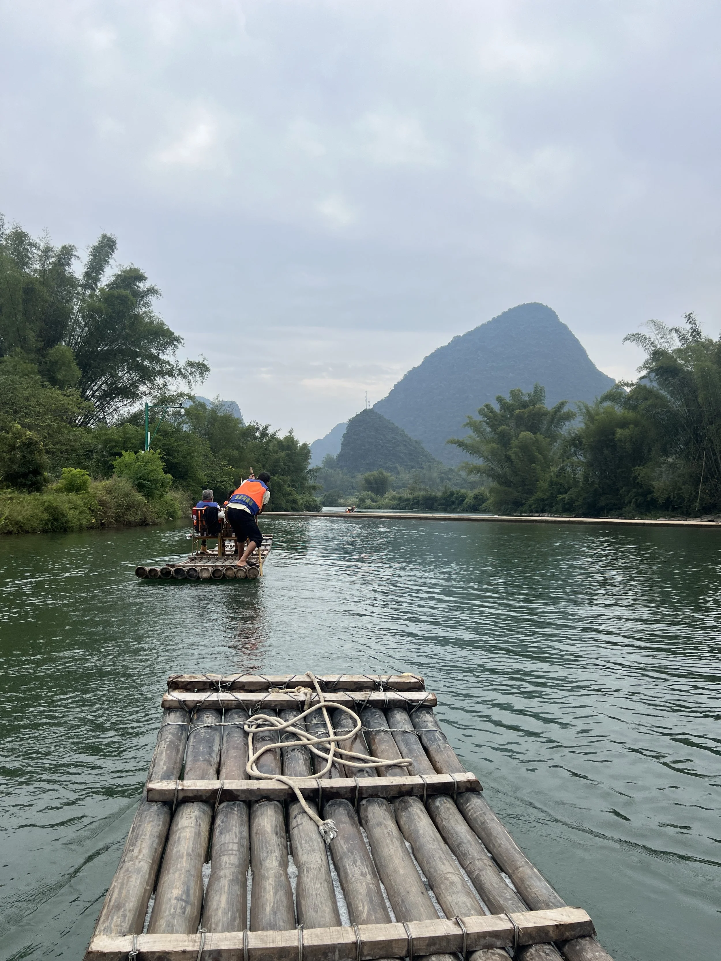 A bamboo raft with two passengers being poled along a calm green river surrounded by lush karst limestone peaks in the Guilin countryside, Guangxi, China