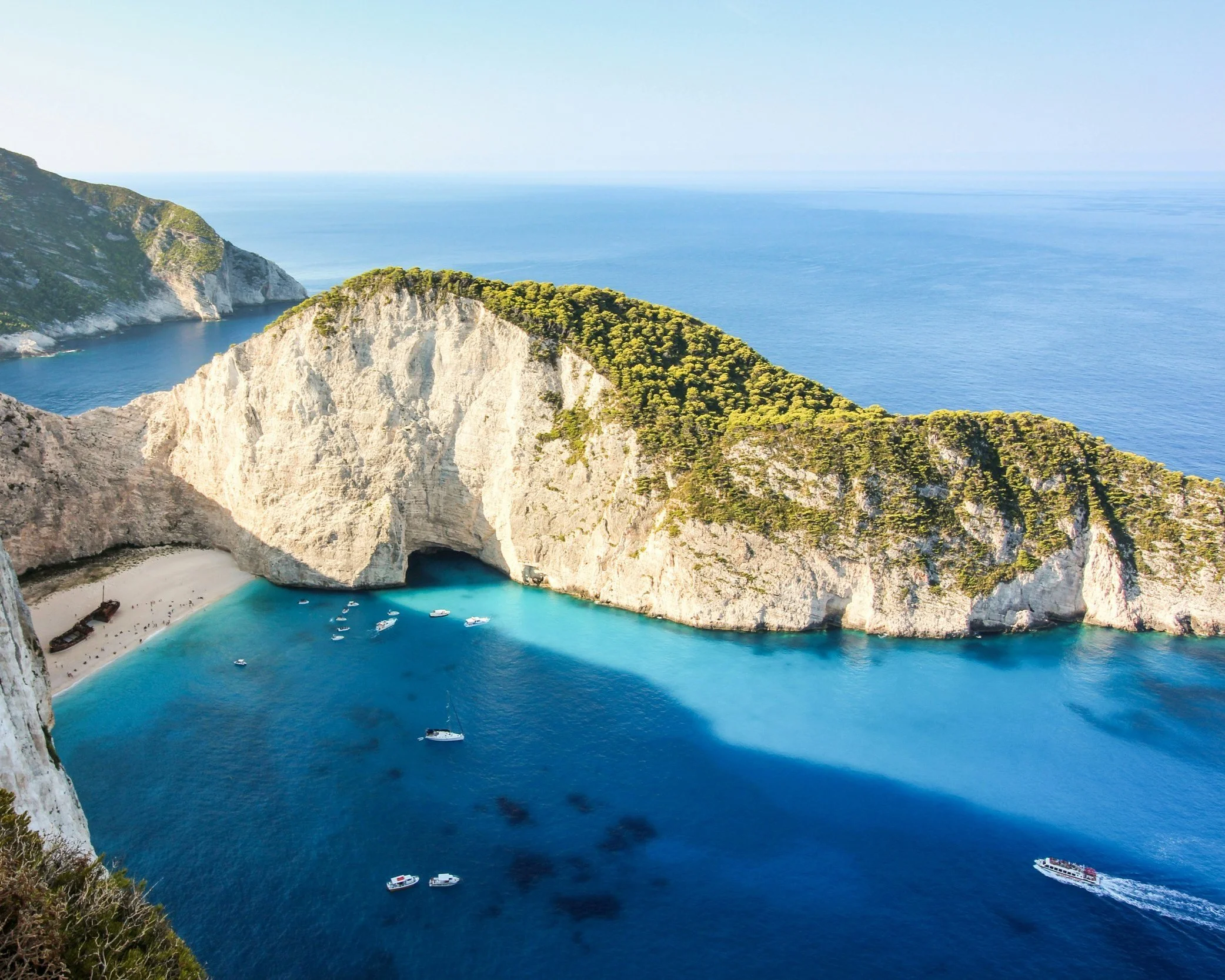 Aerial view of the famous Navagio shipwreck cove on Zakynthos, with towering white limestone cliffs, vivid turquoise water and boats anchored in the enclosed bay, Greece