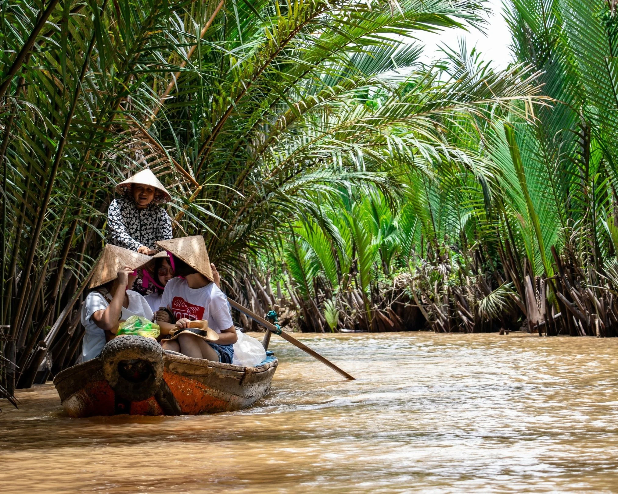A woman in a conical hat rowing a wooden boat with a passenger through a narrow waterway densely lined with nipa palm trees along the Mekong River in Cambodia