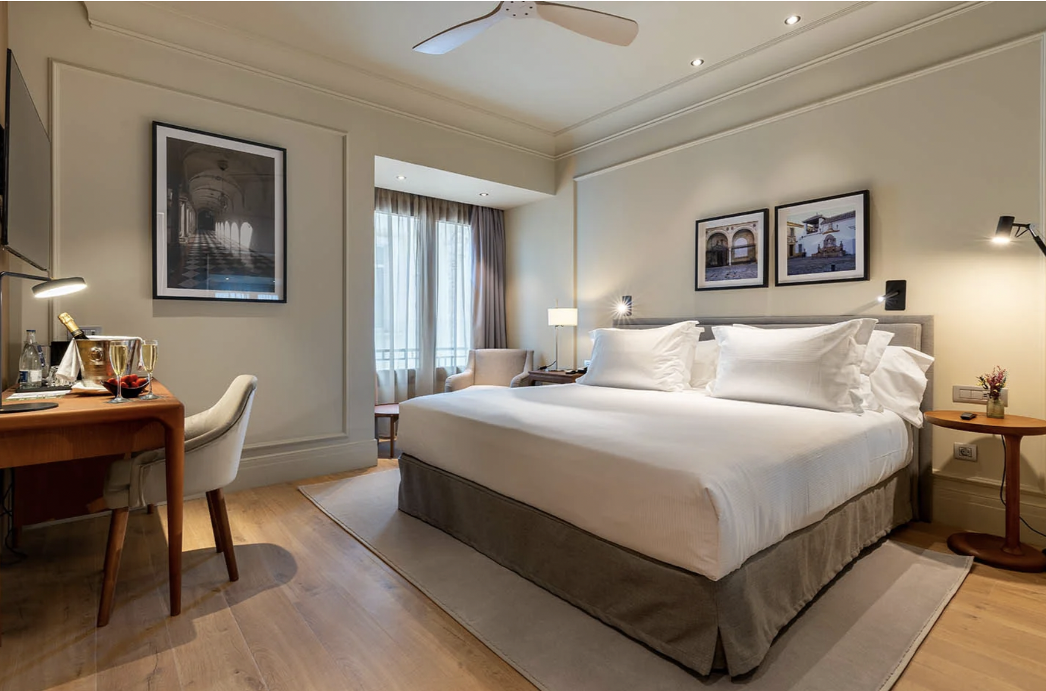 A well-lit hotel room with a large bed with white linens, two framed pictures above it, a small round side table with a flower vase, a chair by the window, and a desk with glasses and a bottle. The room has light-colored walls, wood flooring, and a ceiling fan.