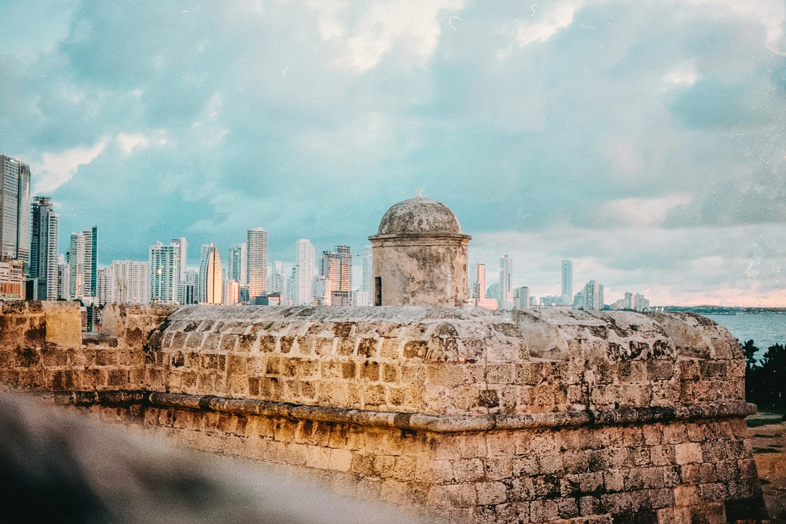 Stone watchtower turret on the 16th-century city walls of Cartagena contrasting with the modern glass skyscraper skyline of Bocagrande in the background at dusk, Colombia