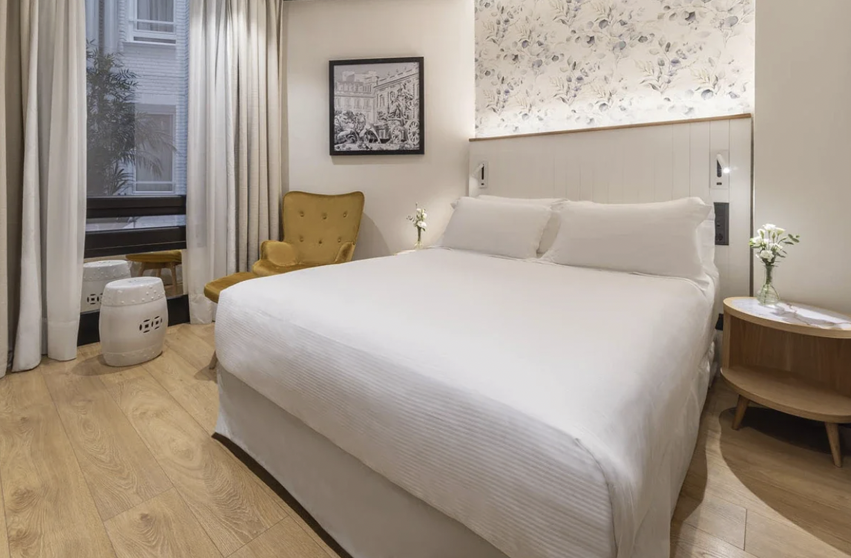 Cozy hotel room with a white bed, mustard-colored armchair, side table with flowers, large window with curtains, and framed wall art.