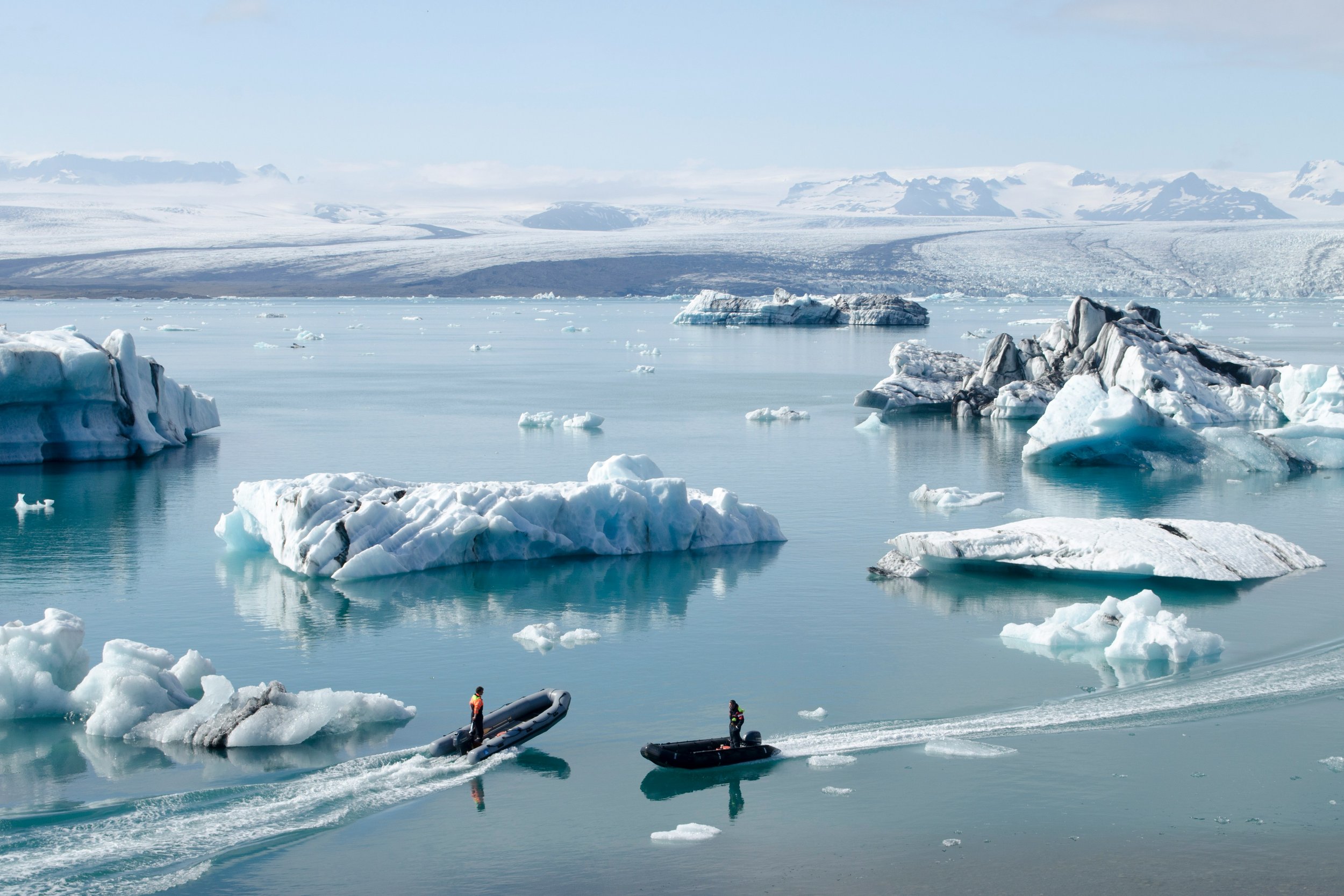 A small zodiac boat navigating between floating icebergs on the Jökulsárlón glacier lagoon in southeast Iceland, with the vast Vatnajökull ice cap in the background
