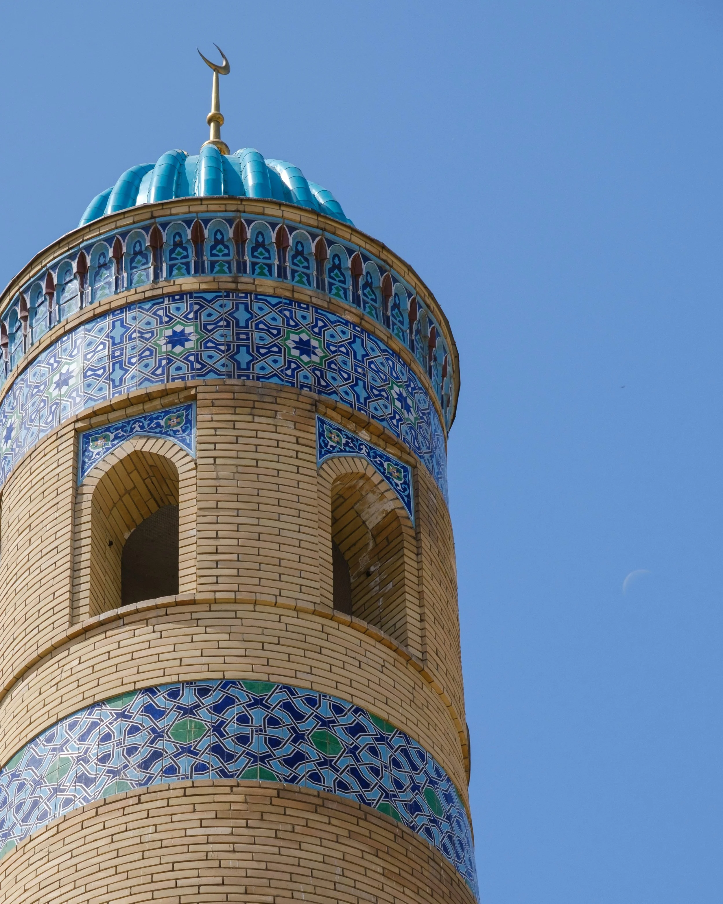 Close-up of a mosque's minaret showcasing intricate blue, green, and white tile patterns against a clear blue sky with a visible crescent moon.