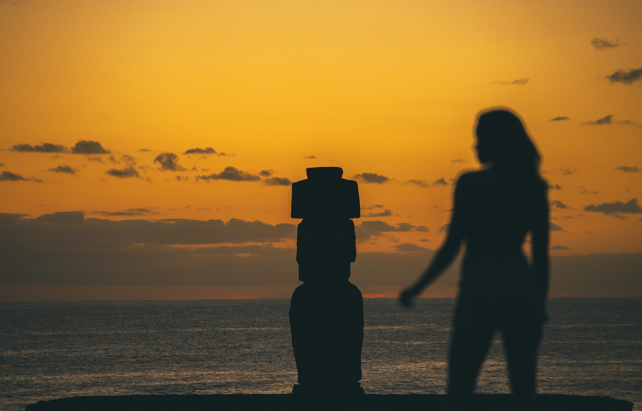 A person silhouetted against a vivid orange sunset sky beside a lone Moai statue on Easter Island, Rapa Nui, Chile