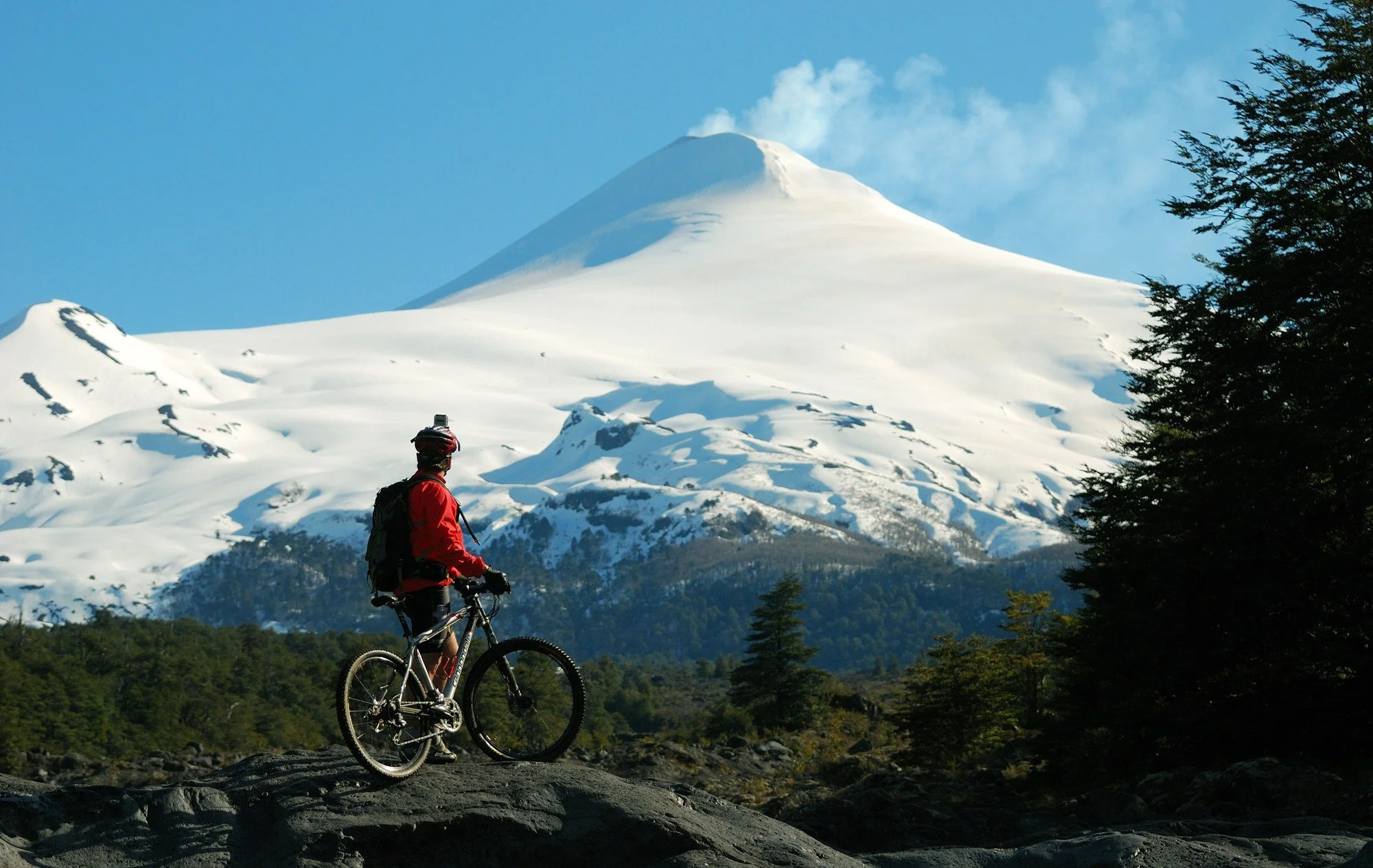 Mountain biker resting on rocks above the treeline with the snow-covered cone of Villarrica volcano behind them in Pucón, Chile