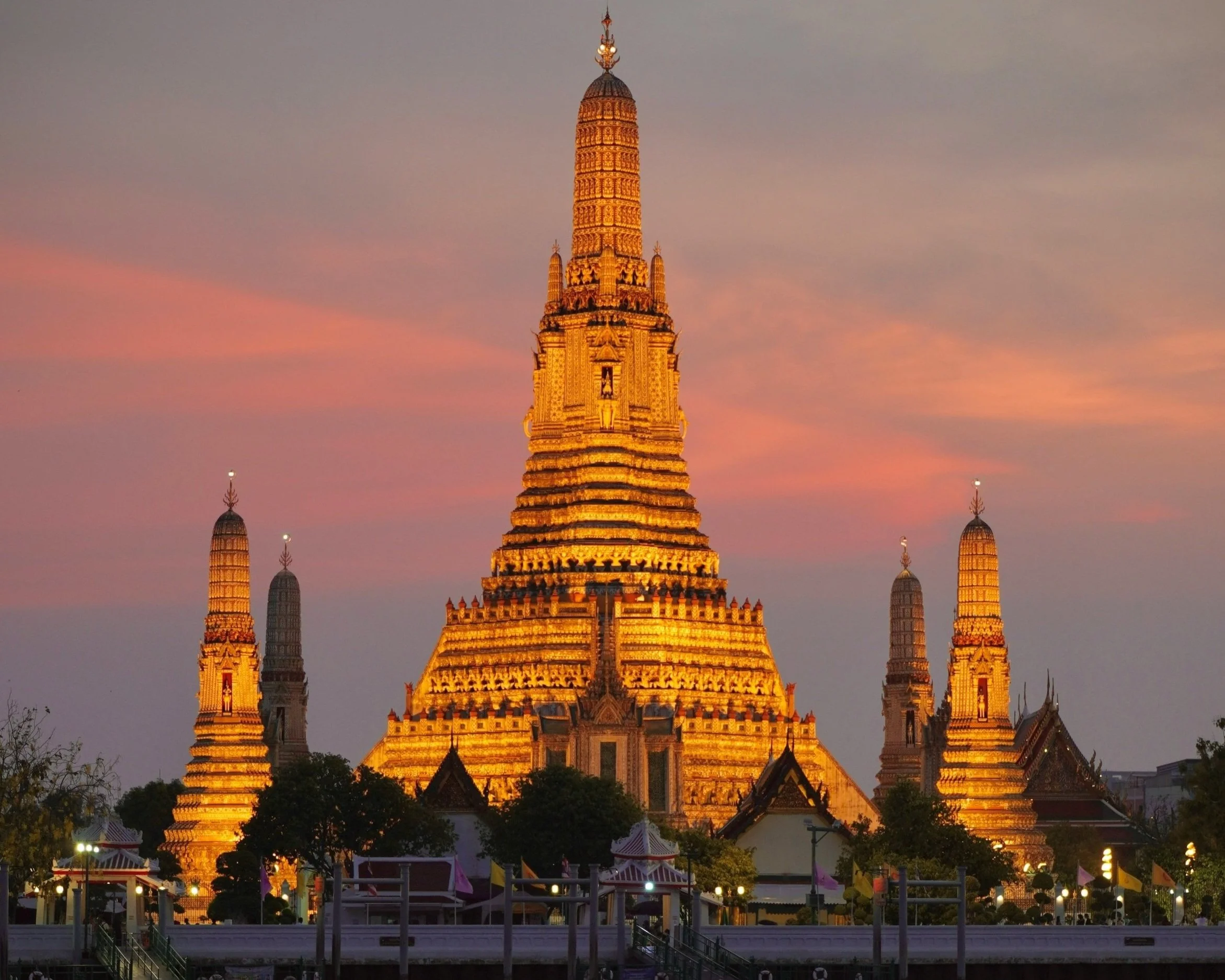 Wat Arun temple complex glowing gold against a pink and purple sunset sky in Bangkok, Thailand, with the central prang tower and four smaller satellite towers reflected in warm evening light