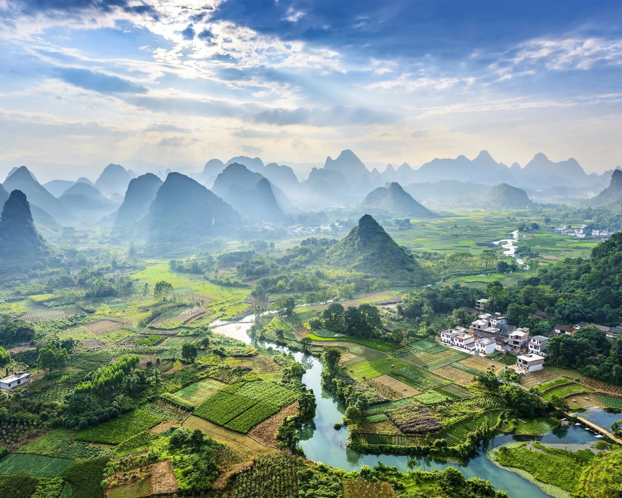 Aerial view of the Guilin karst landscape in Guangxi, China, showing dramatic limestone peaks rising above a patchwork of green rice fields, a winding river and small villages under a partly cloudy sky