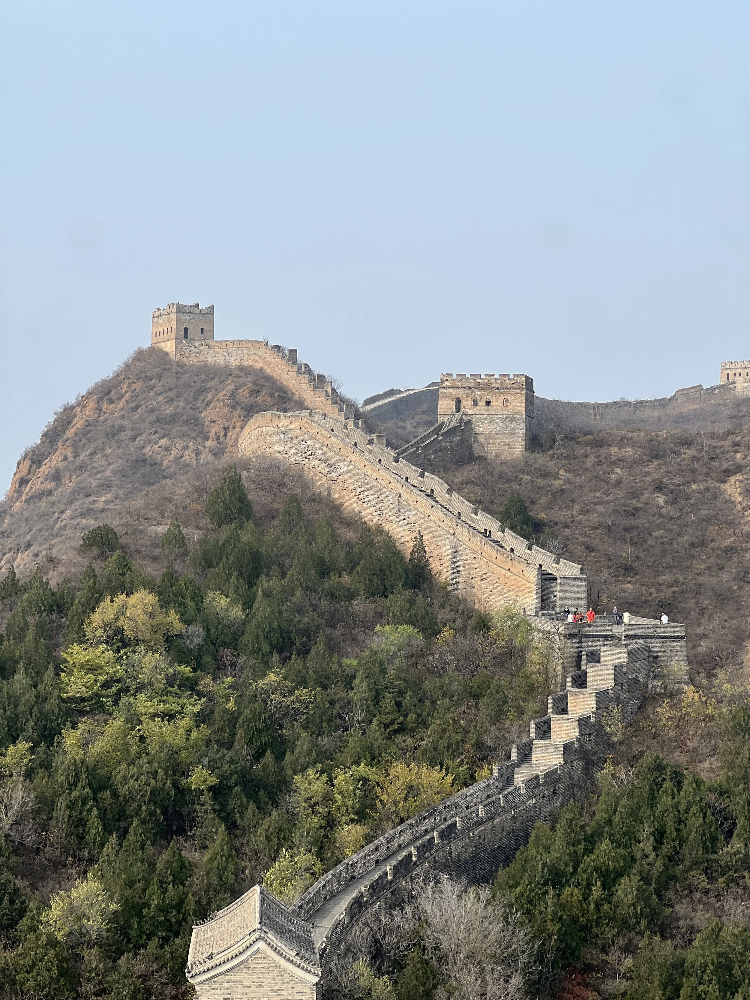 A restored section of the Great Wall of China stretching over steep barren hills near Beijing, with watchtowers visible along the battlements and a clear pale sky above