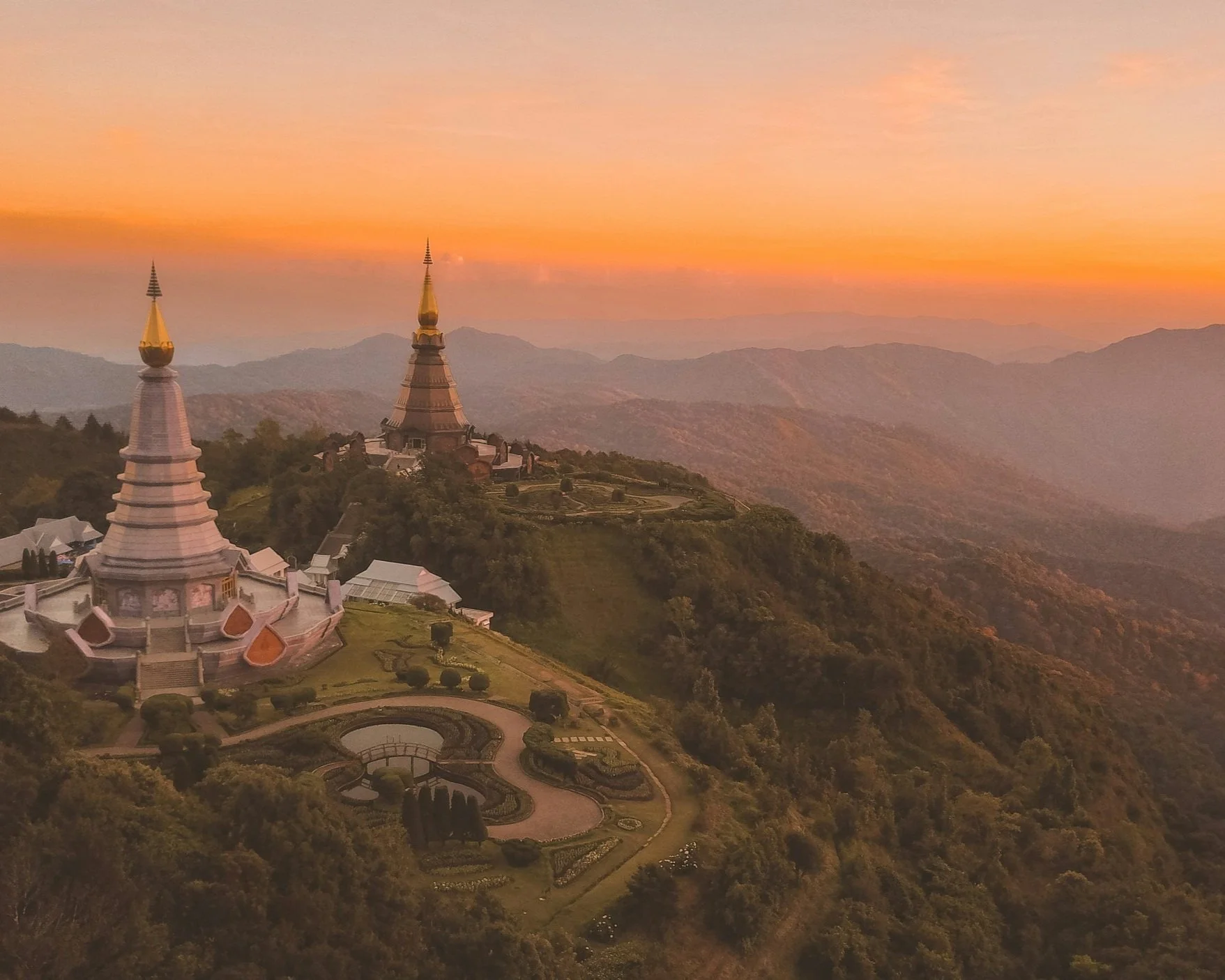 Aerial view of the twin white chedis of Doi Inthanon National Park at sunset, surrounded by forested ridgelines and layered mountain haze in northern Thailand near Chiang Mai