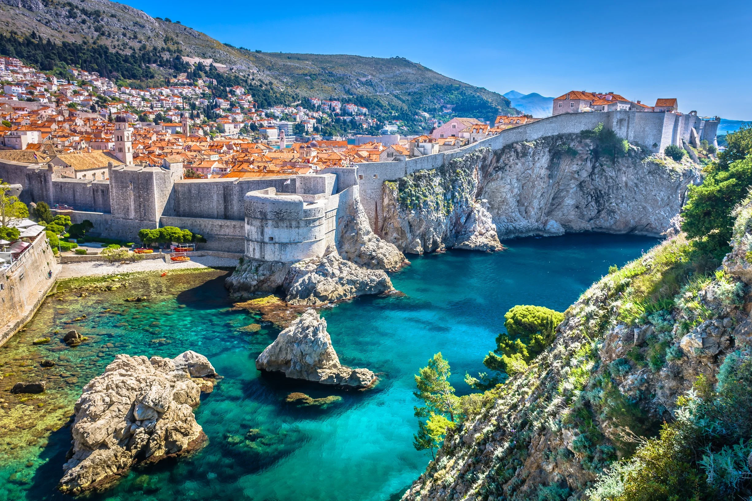 Dubrovnik's imposing medieval city walls curving along a rocky limestone coastline above a turquoise Adriatic cove, with the old town rooftops and mountains beyond, Croatia