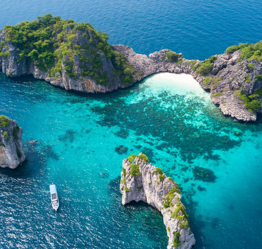 Aerial view of rocky islands with lush green vegetation surrounded by turquoise water, featuring a small boat near the rocks, and a white sand beach in the background.