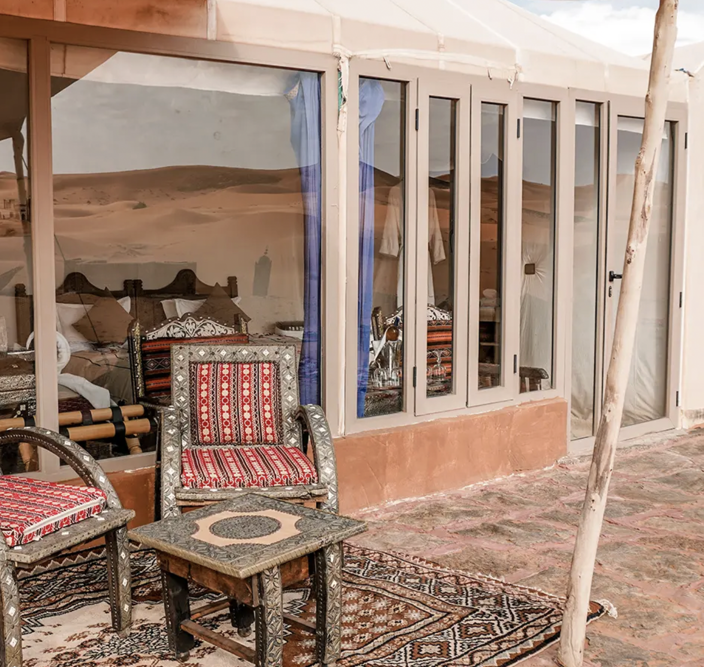 A room with large glass windows and desert landscape reflections. Decor includes patterned chairs, small tables, and a rug, with sandy walls and a brick floor.