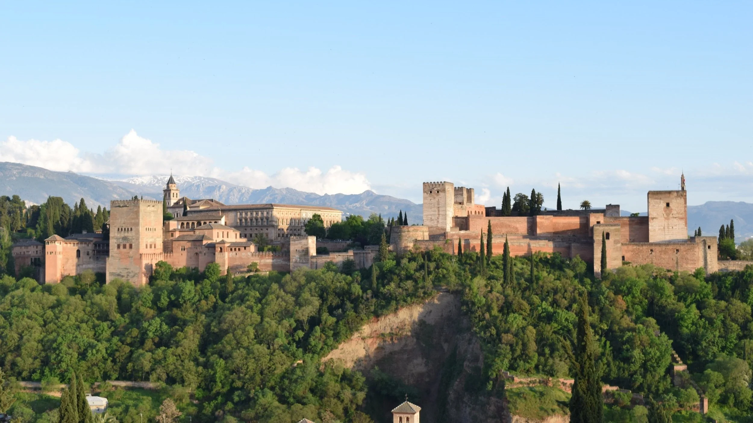 Panoramic view of the Alhambra palace and fortress complex rising above the forests of Granada, with the Sierra Nevada mountains behind