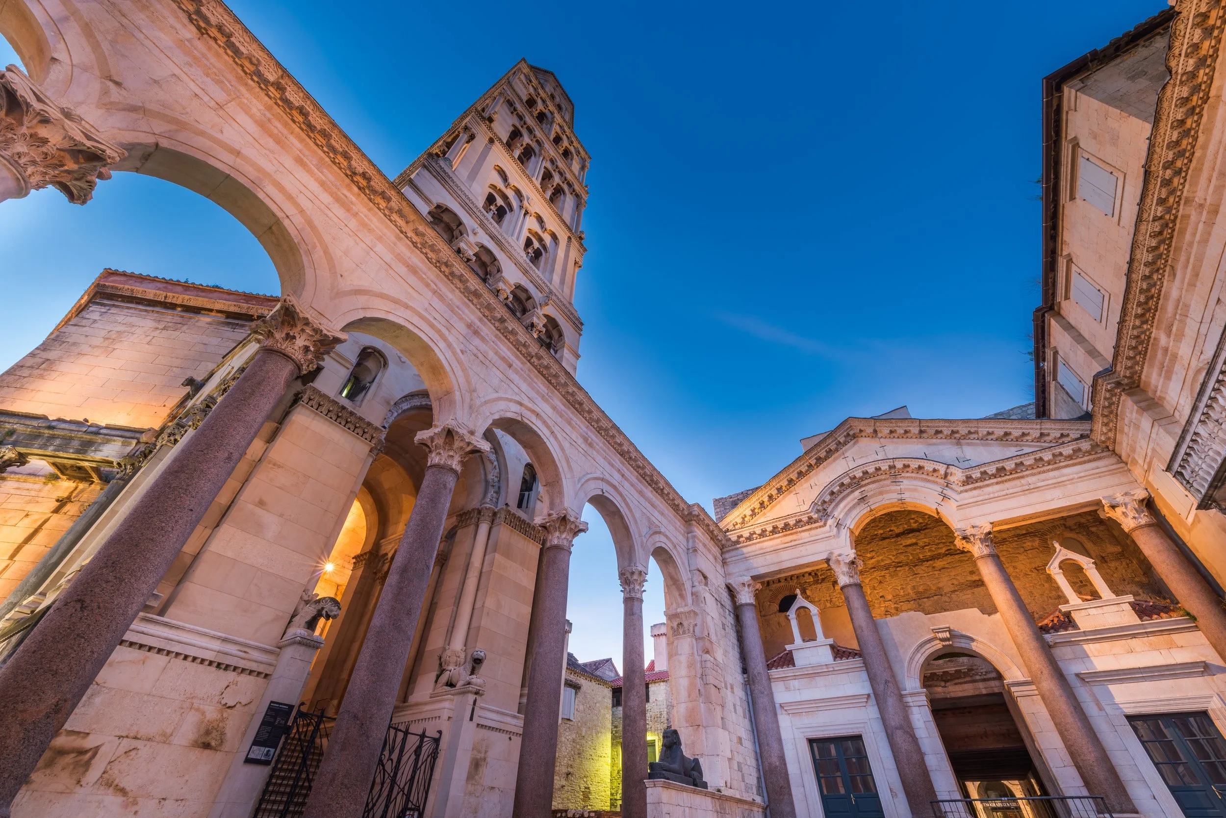 Looking up through the ancient Roman arches of the Diocletian's Palace peristyle in Split, Croatia, with the cathedral bell tower rising against a deep blue sky