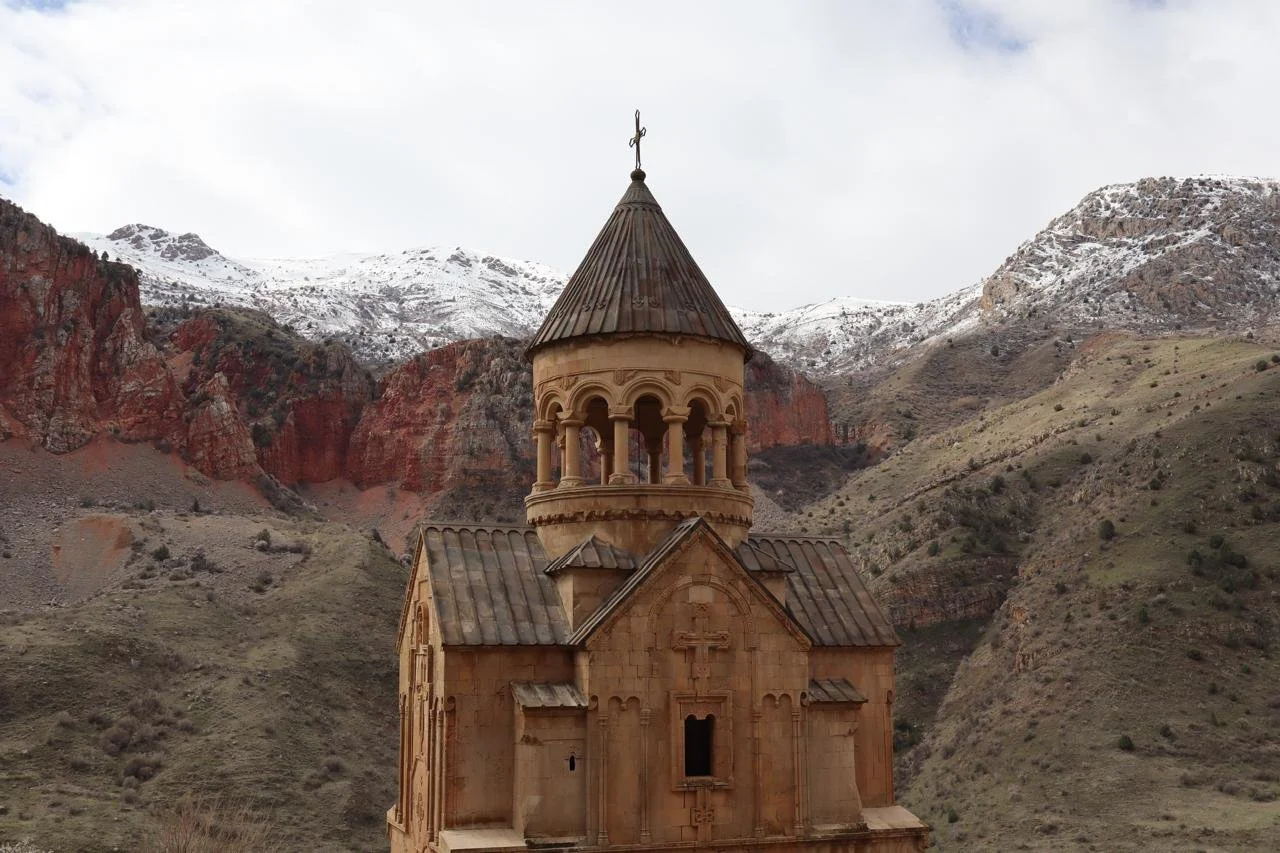 Close frontal view of Noravank monastery church with its conical drum bell tower and carved stone cross facade, set directly against dramatic red sandstone canyon walls and snow-dusted peaks in Vayots Dzor, Armenia