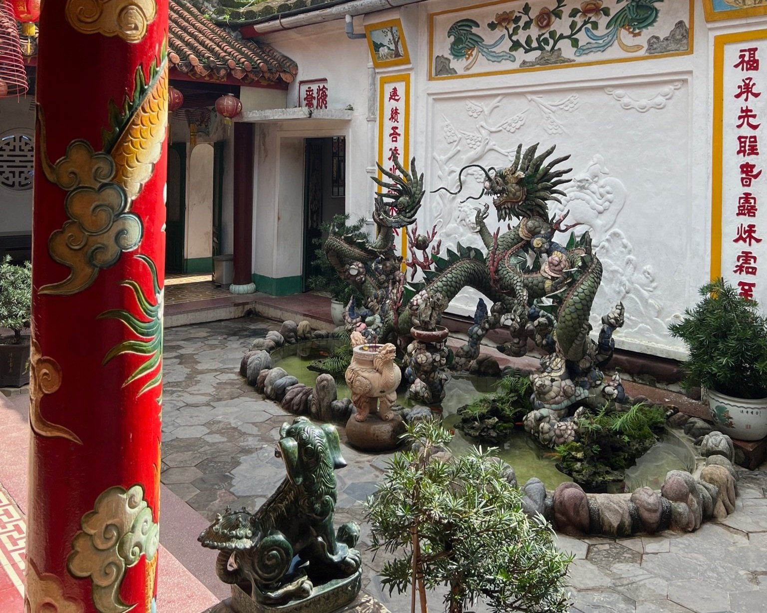 Ornate dragon sculpture and bonsai plants in the courtyard of a traditional Vietnamese assembly hall temple in Hoi An, with red lacquered pillars and carved decorative walls visible behind