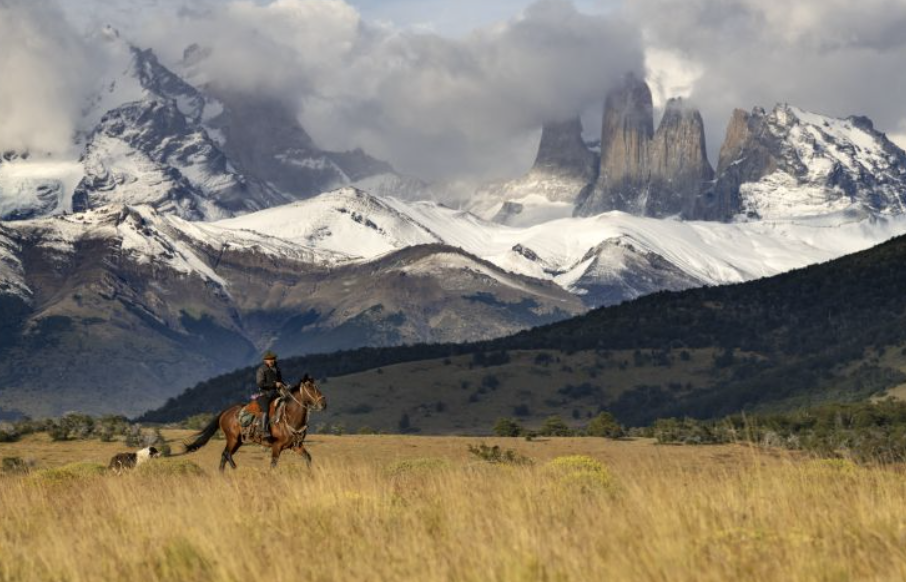 A gaucho on horseback riding across the golden Patagonian steppe with the snow-covered granite towers of Torres del Paine rising dramatically in the background, Chile