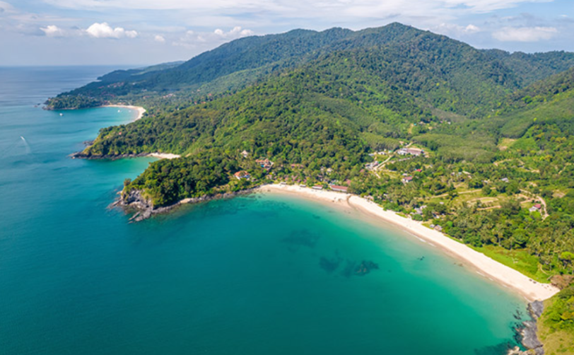 Aerial view of Koh Lanta island showing a curved sandy beach, turquoise shallow water and dense green forest covering the hillside above the coastline in the Andaman Sea, Thailand
