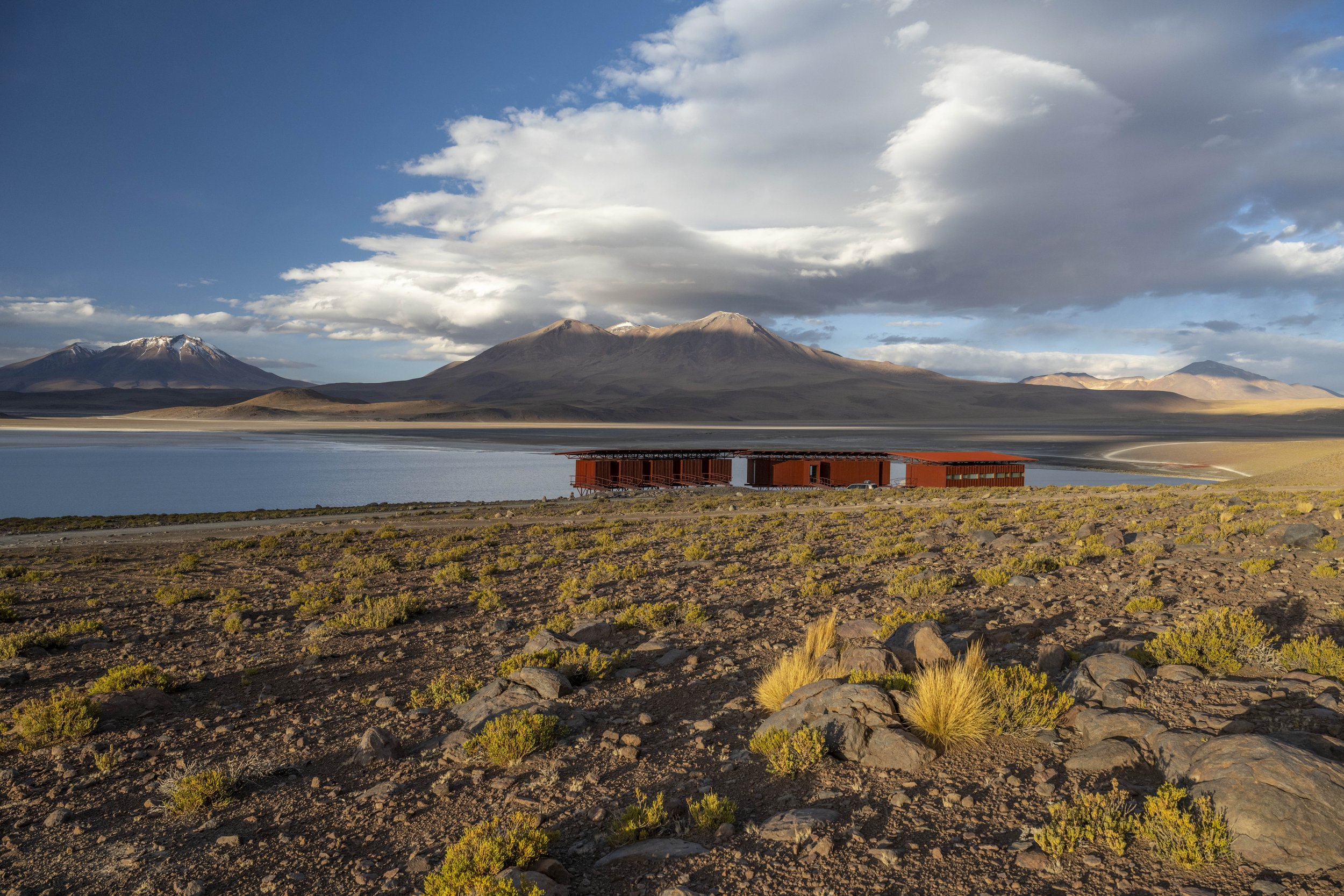A scenic landscape with desert terrain in the foreground, a large lake in the middle ground, and snow-capped mountains and a partly cloudy sky in the background.