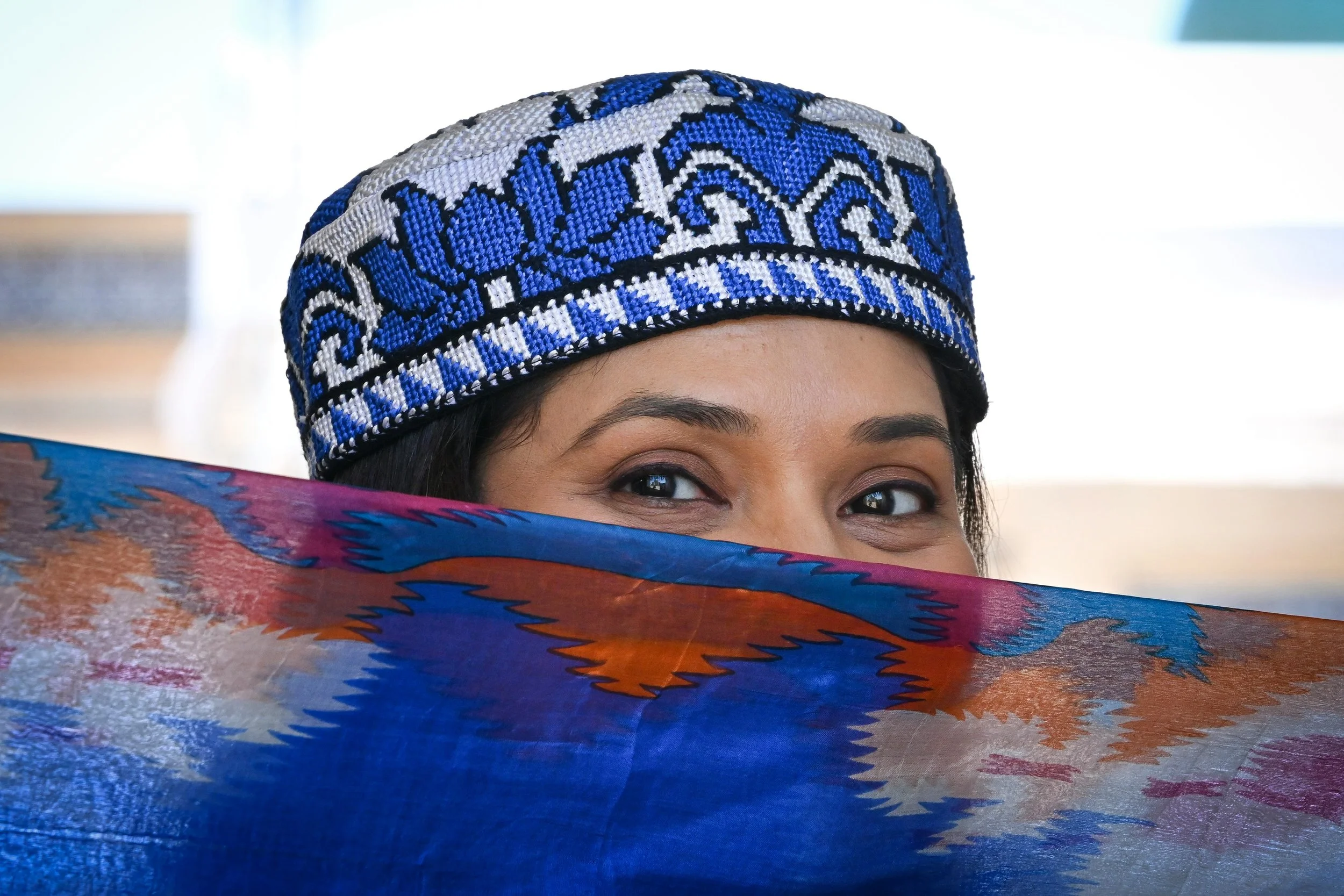 Woman wearing an embroidered blue and black cap peeking over a colorful fabric with orange, blue, and pink patterns.