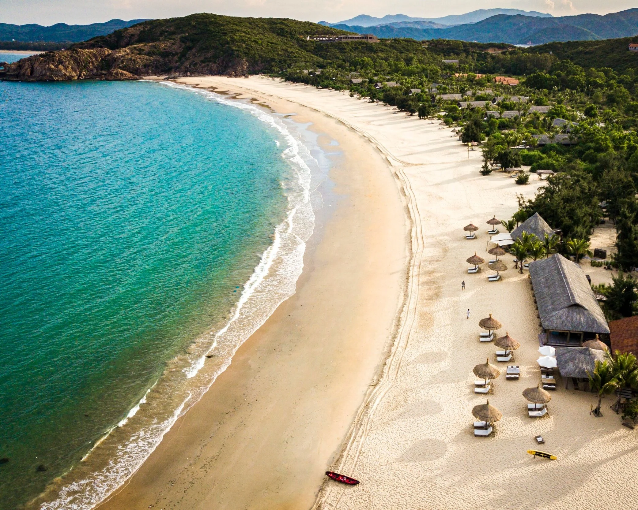 Aerial view of a curved white sand beach in Nha Trang, Vietnam, with shallow turquoise water, a row of beach umbrellas and a forested mountain headland visible in the background