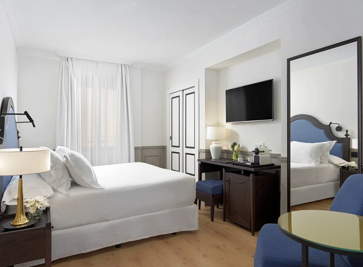 A hotel room with a white bed, bedside lamp, TV, mirror, desk, small chair, and a blue chair with a round table.
