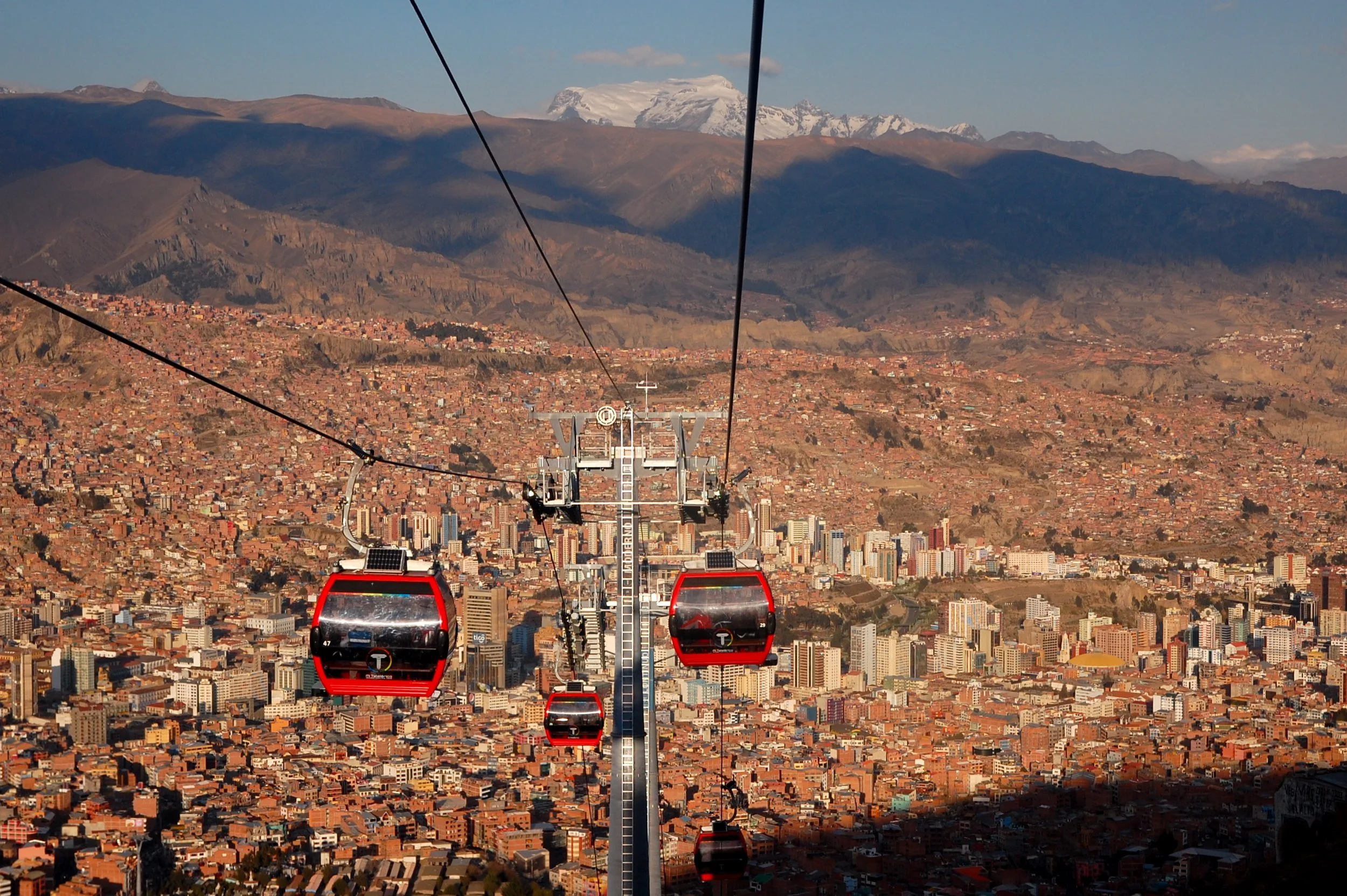 Red cable car gondolas floating above the dense urban sprawl of La Paz with snow-capped Andean peaks rising in the distance, Bolivia