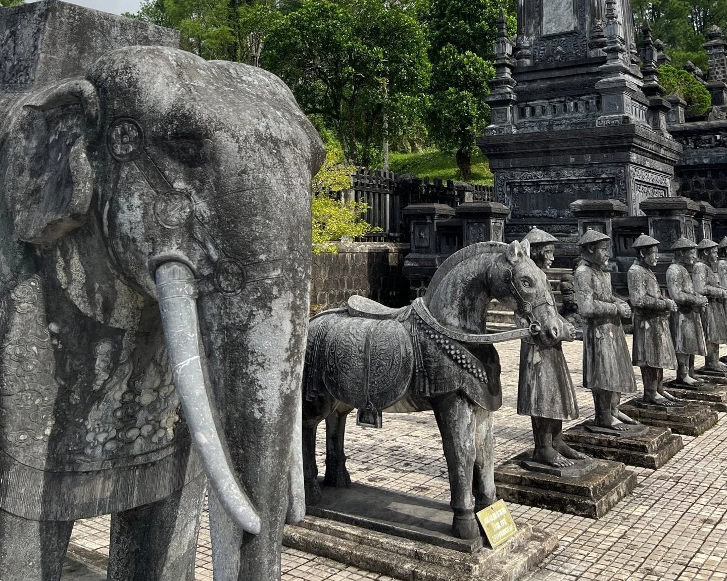Stone elephant and horse statues alongside carved mandarin guardian figures lining the courtyard of a Nguyen dynasty imperial tomb in Hue, Vietnam, surrounded by tropical greenery
