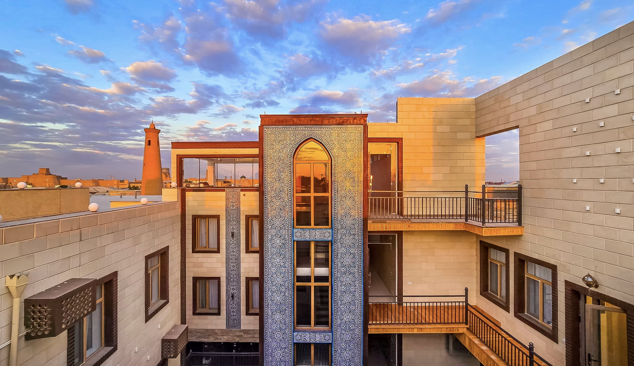 Sunset view of a rooftop hotel with beige brick walls, wooden balconies, and a decorative blue-tiled archway in a Middle Eastern style.