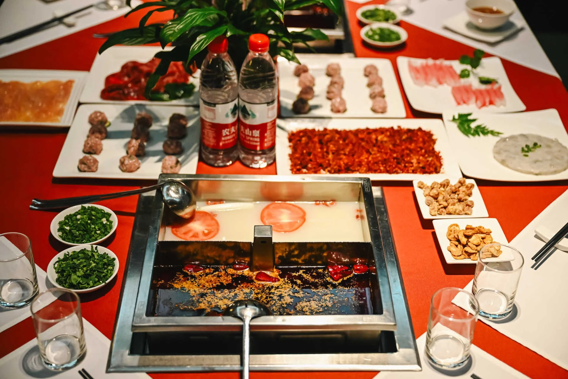 A Sichuan hot pot table in Chengdu with a divided broth pot, surrounded by plates of raw meat, vegetables, dried chillies and condiments ready to cook, with a bottle of water and chopsticks on a red tablecloth