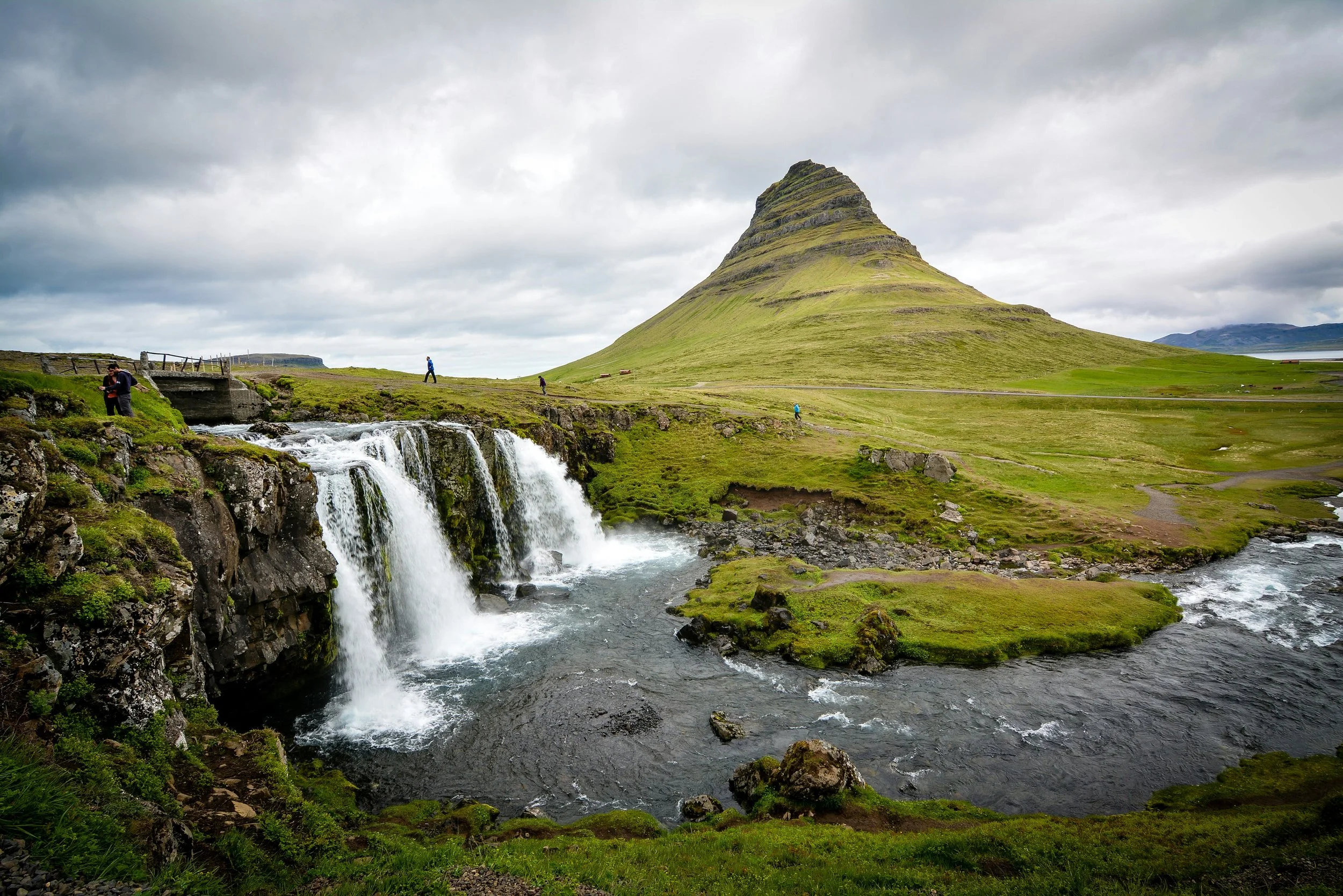 The iconic cone-shaped Kirkjufell mountain rising above a cascading waterfall and lush green meadows in the Snæfellsnes Peninsula, northern Iceland